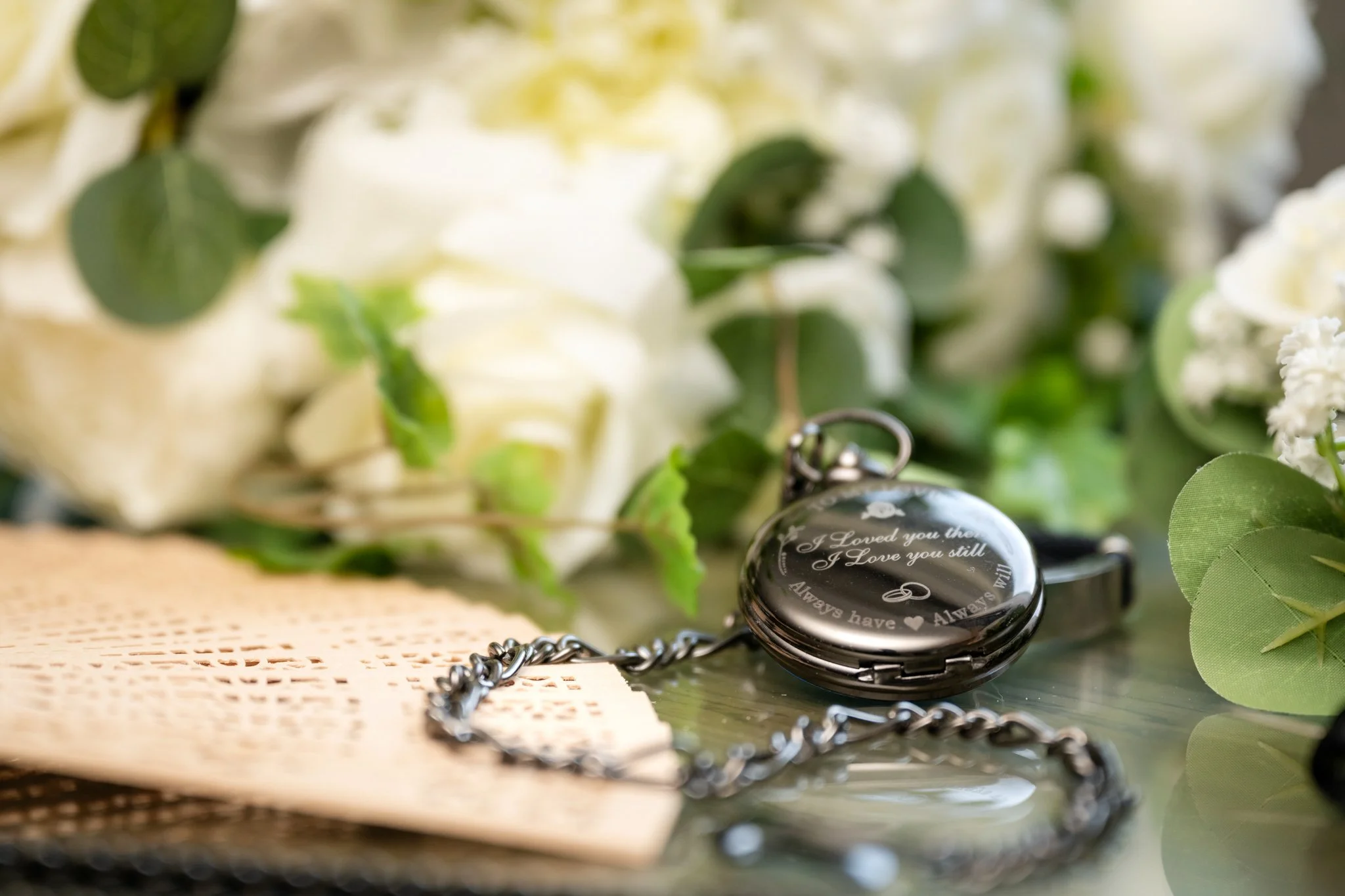 A pocket watch with engraved romantic message, surrounded by white flowers and green leaves.