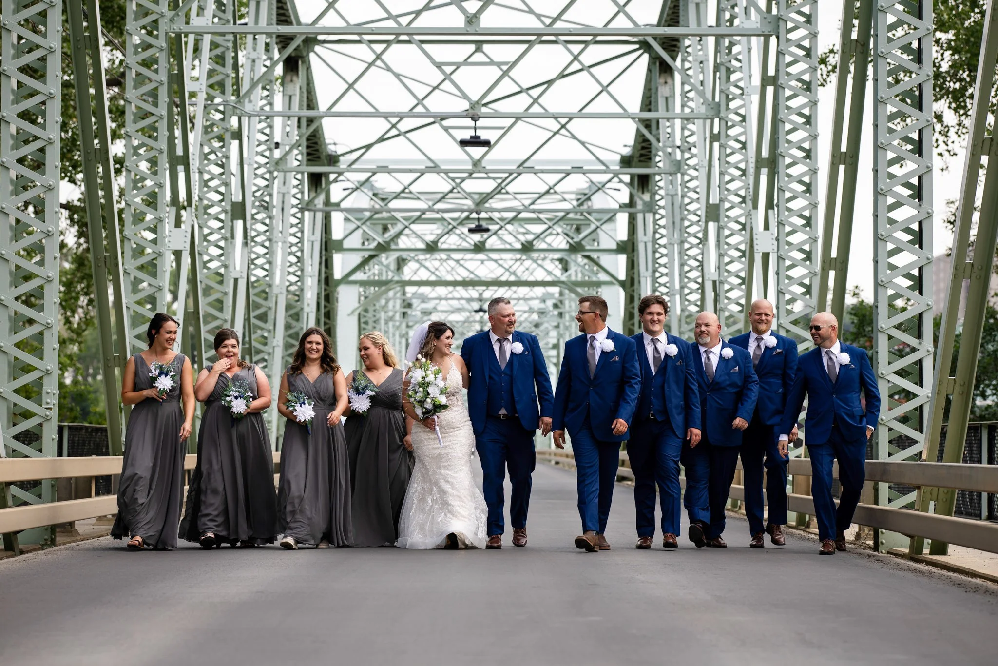 A wedding party walking across a metal bridge, with the bride and groom in the center, surrounded by bridesmaids and groomsmen in formal attire.