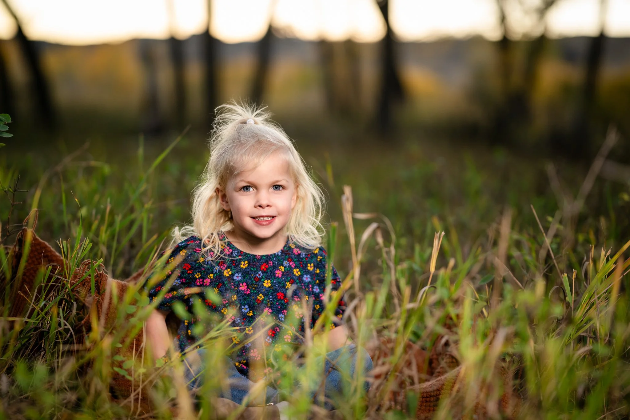 Young girl with blonde curly hair wearing a floral shirt, sitting outdoors in tall grass with blurred trees in the background.