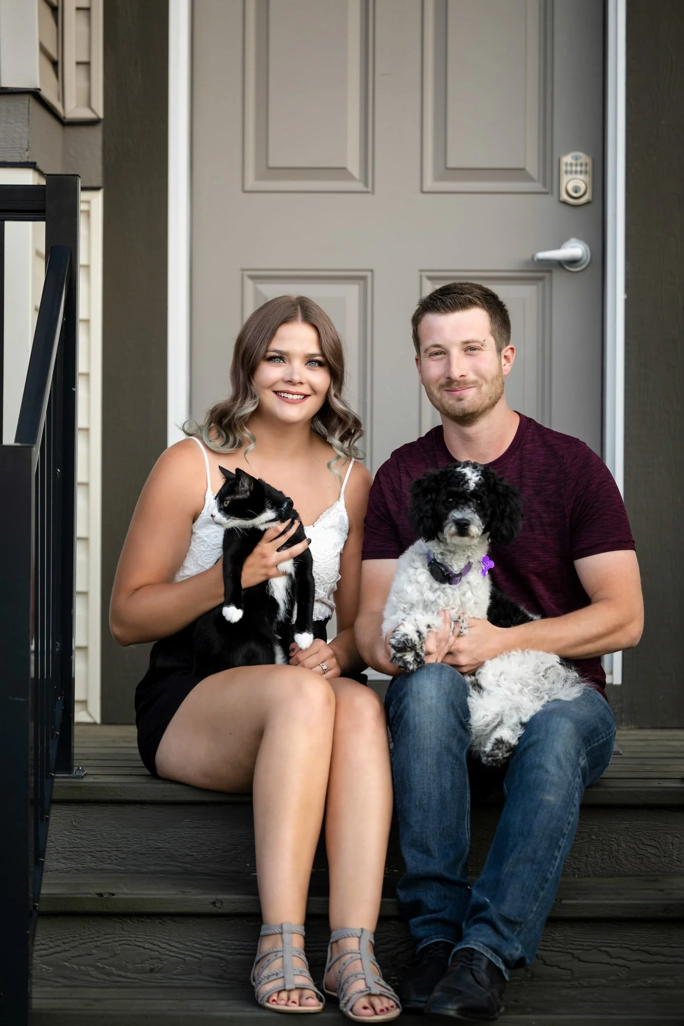 A young woman and a young man sitting on the front steps of a house, holding a black and white cat and a black and white dog, smiling at the camera.