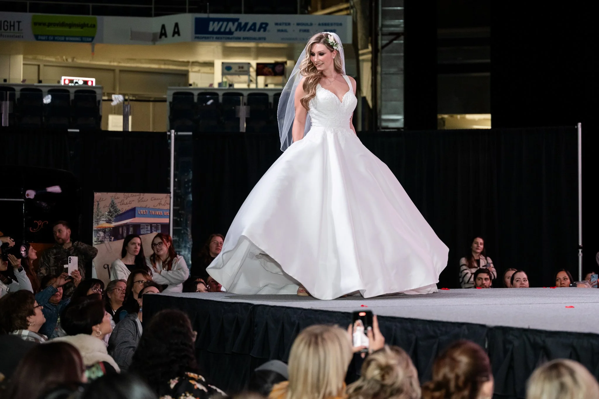 A woman models a wedding dress on a runway with a black curtain backdrop and an audience watching.