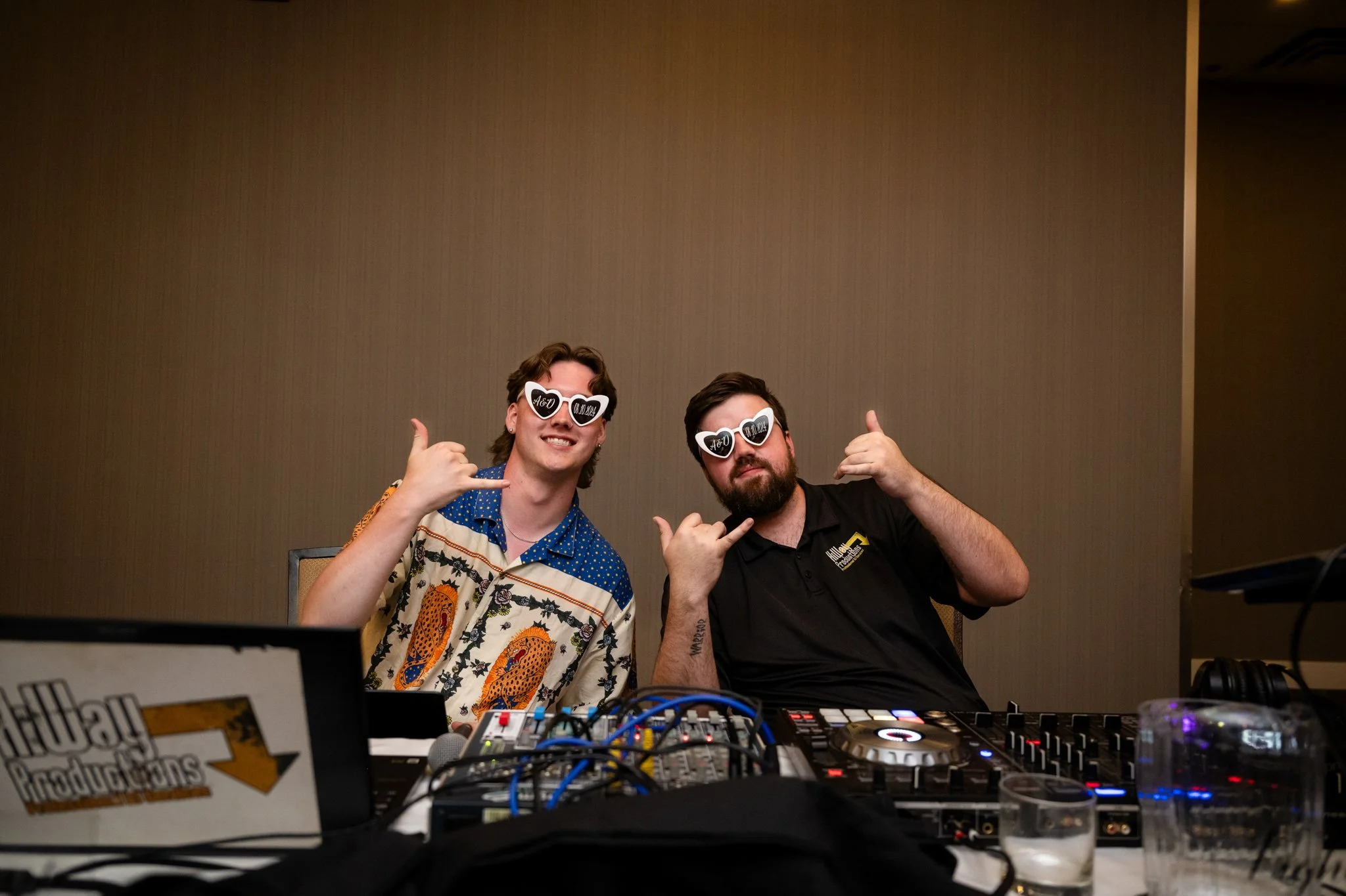 Two young men with long hair and beard, wearing sunglasses with 'You' and 'Me' written on them, posing at a DJ booth with their hands making the 'call me' gesture. One is wearing a colorful patterned shirt, and the other is in a black polo shirt.