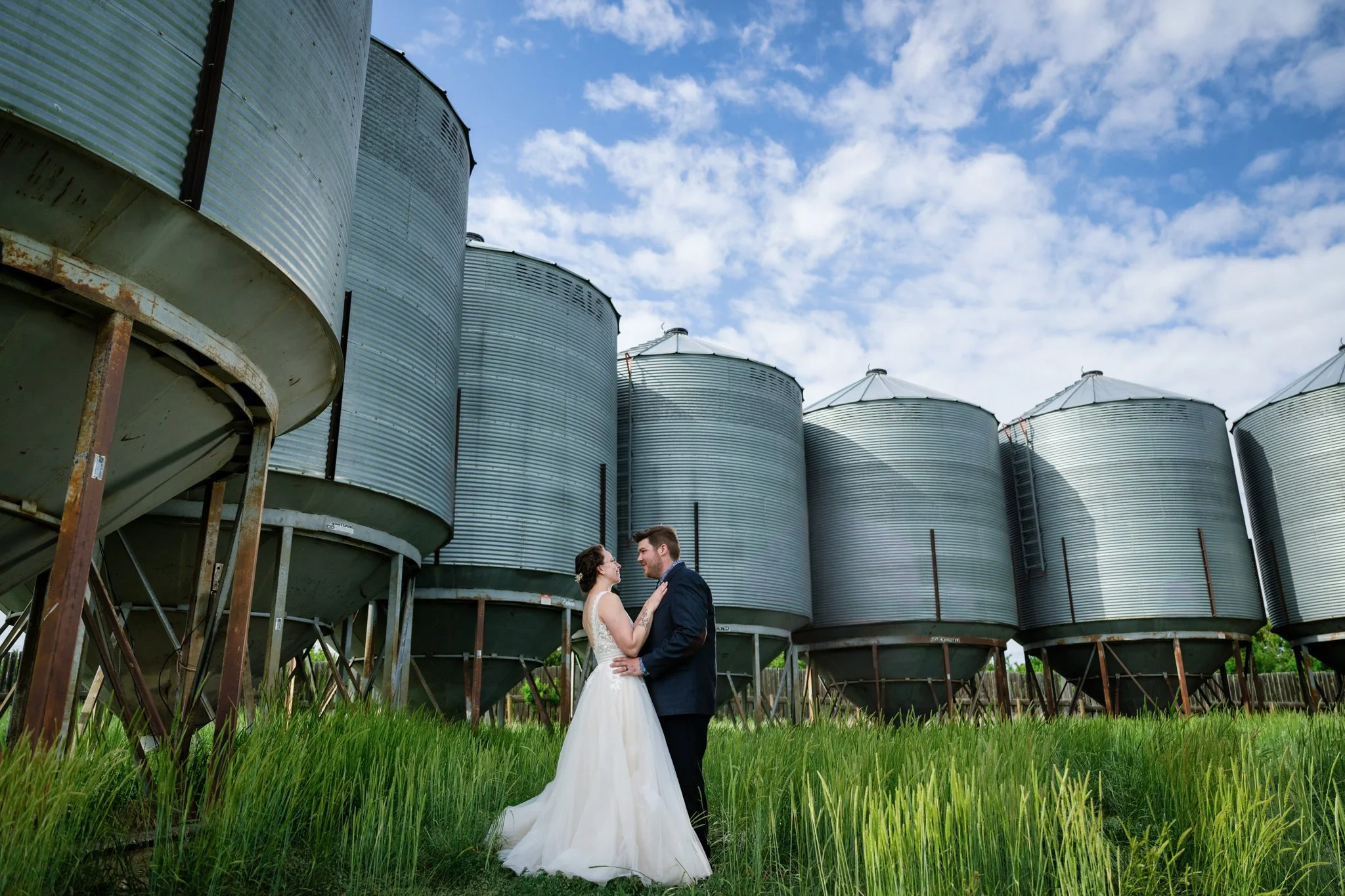 A bride and groom in wedding attire standing close together outdoors, with large metal storage silos and a partly cloudy sky in the background.