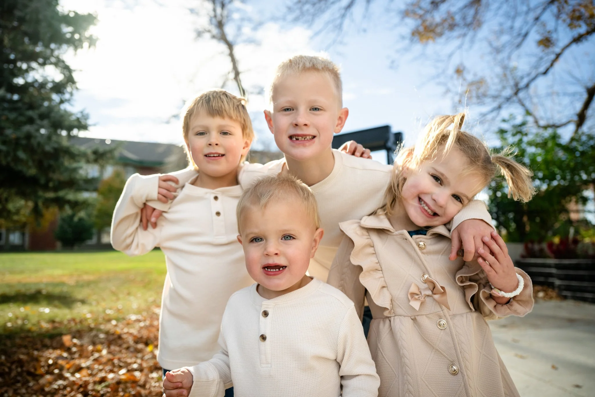 Five children smiling and posing outdoors on a sunny day, with trees and a building in the background.
