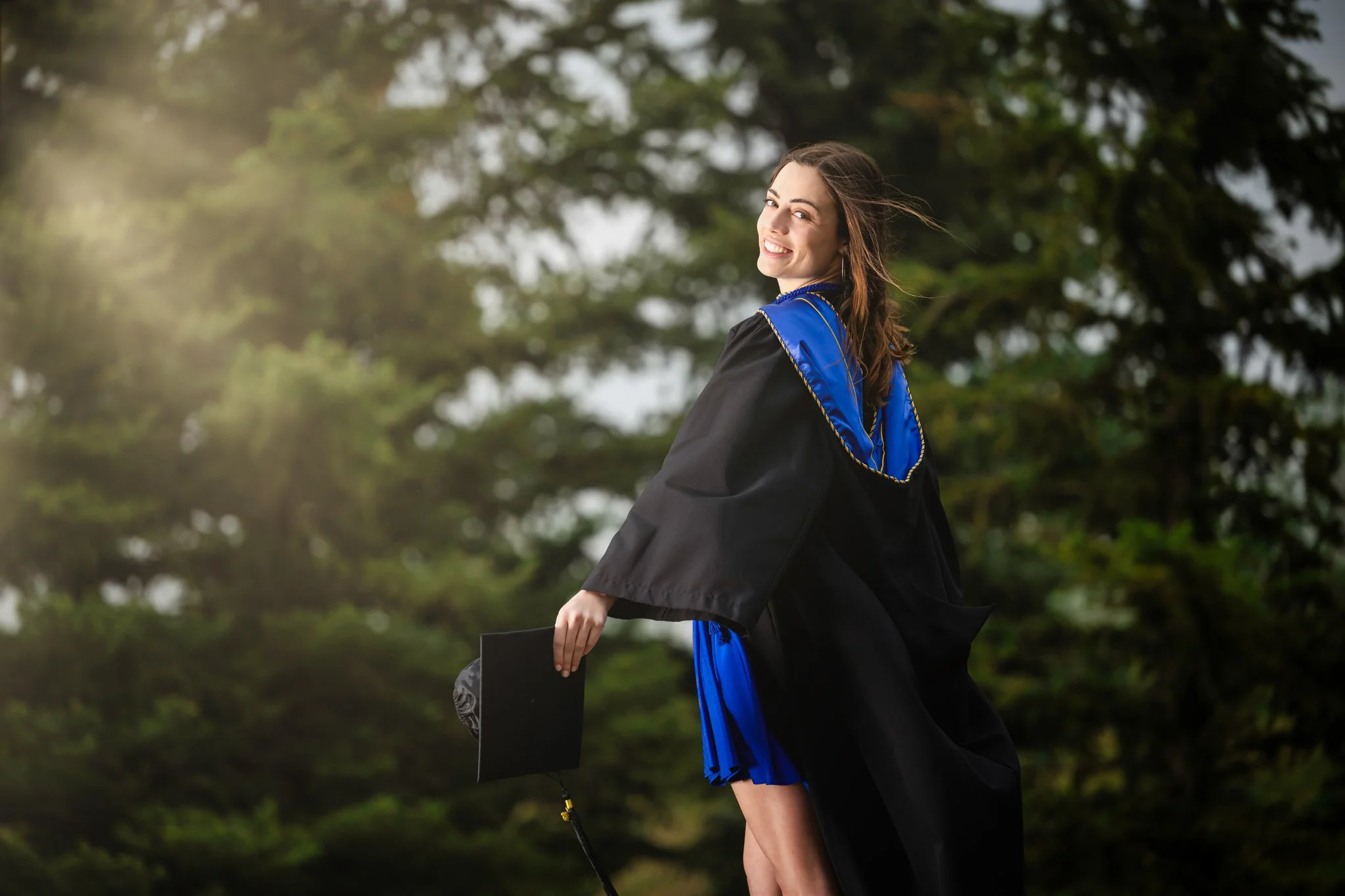 Young woman in graduation cap and gown holding diploma, smiling outdoors with trees in background.