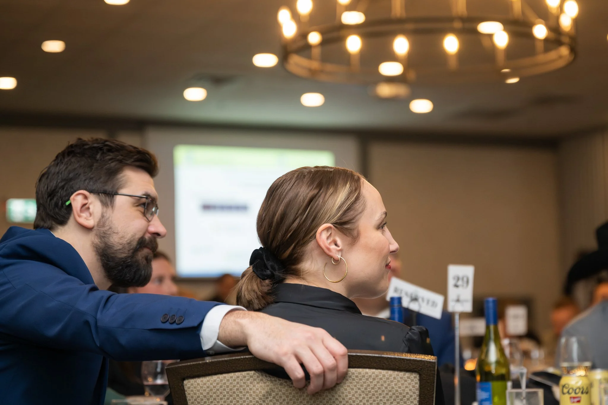 Man in a navy suit and glasses sitting next to a woman with hoop earrings and a black top, at a formal event or conference, with a blurred background of tables, drinks, and event signs.