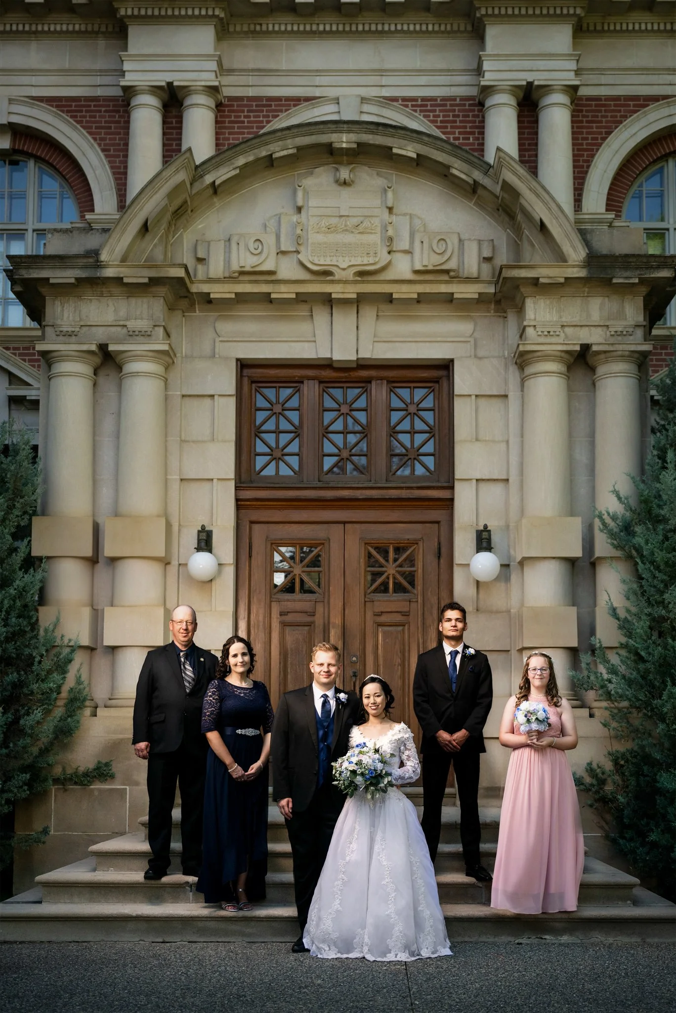 A wedding party standing outside a historic building with a large wooden door and stone architecture. The group includes a bride in a white dress holding a bouquet, groom in a suit, and four other individuals in formal attire, two men and two women.