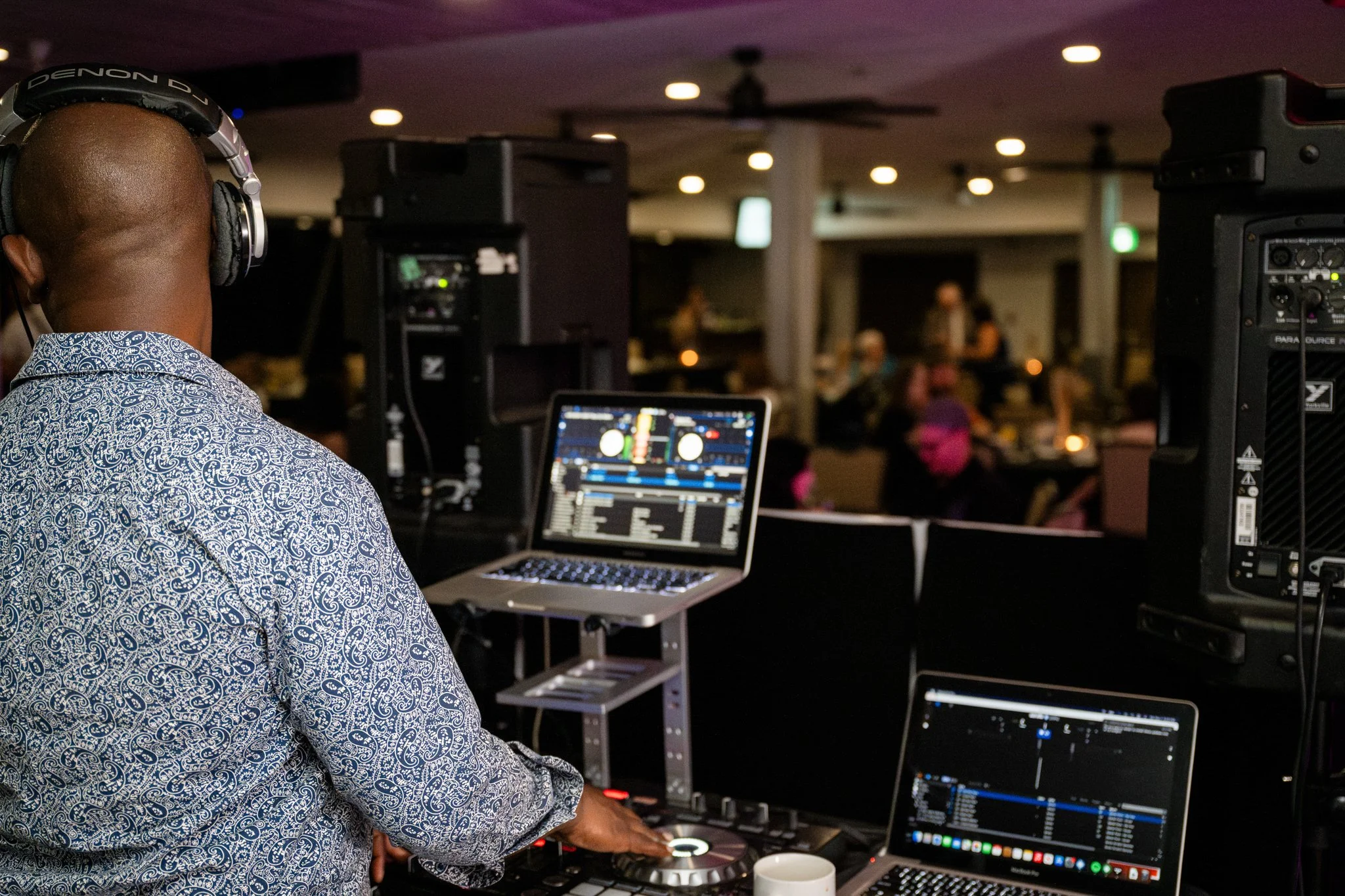 DJ wearing headphones and a patterned shirt operating DJ equipment in a dimly lit nightclub or event space, with a crowd of people in the background.