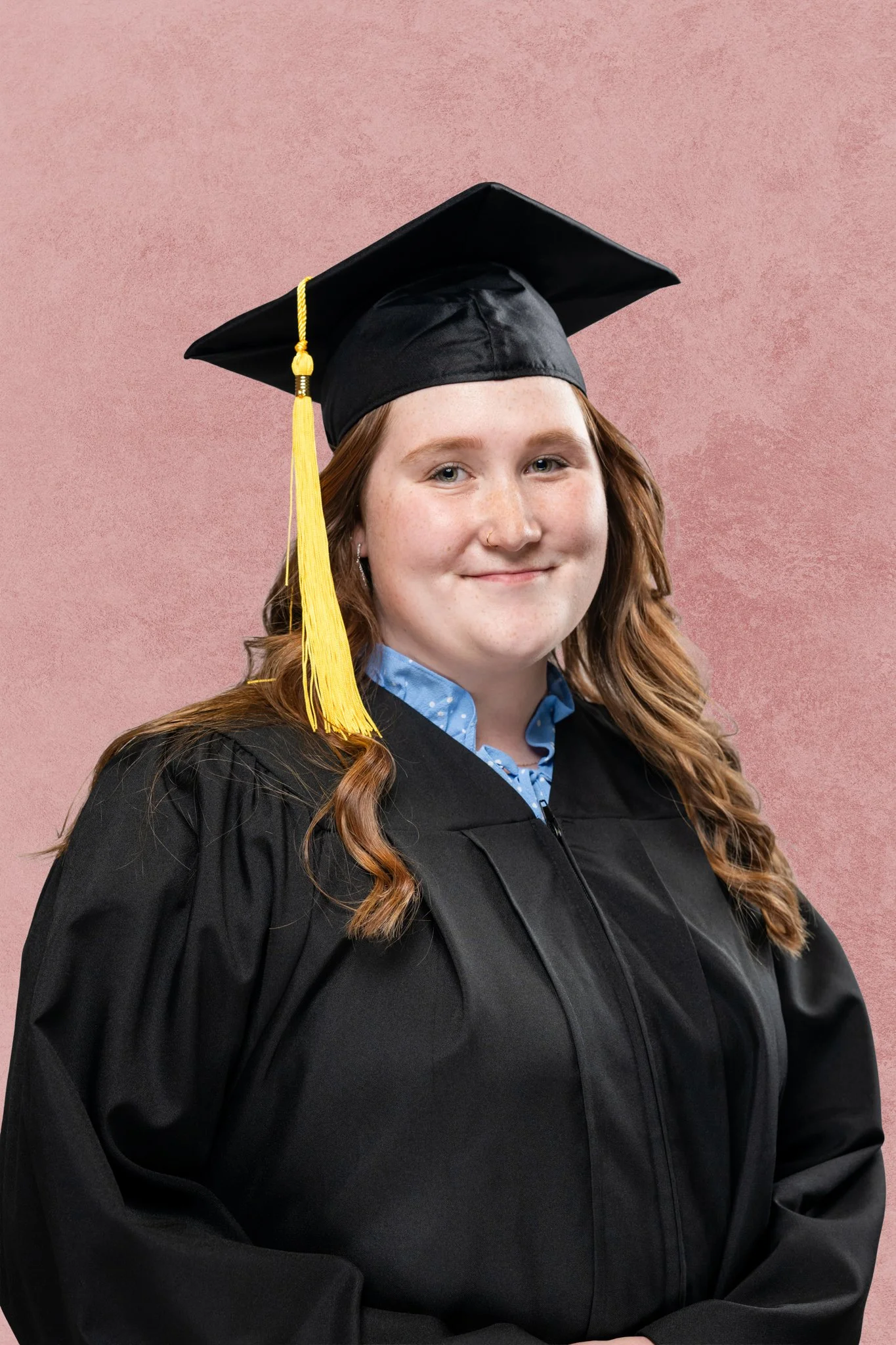 A young woman in graduation cap and gown smiling, standing against a pink textured background.