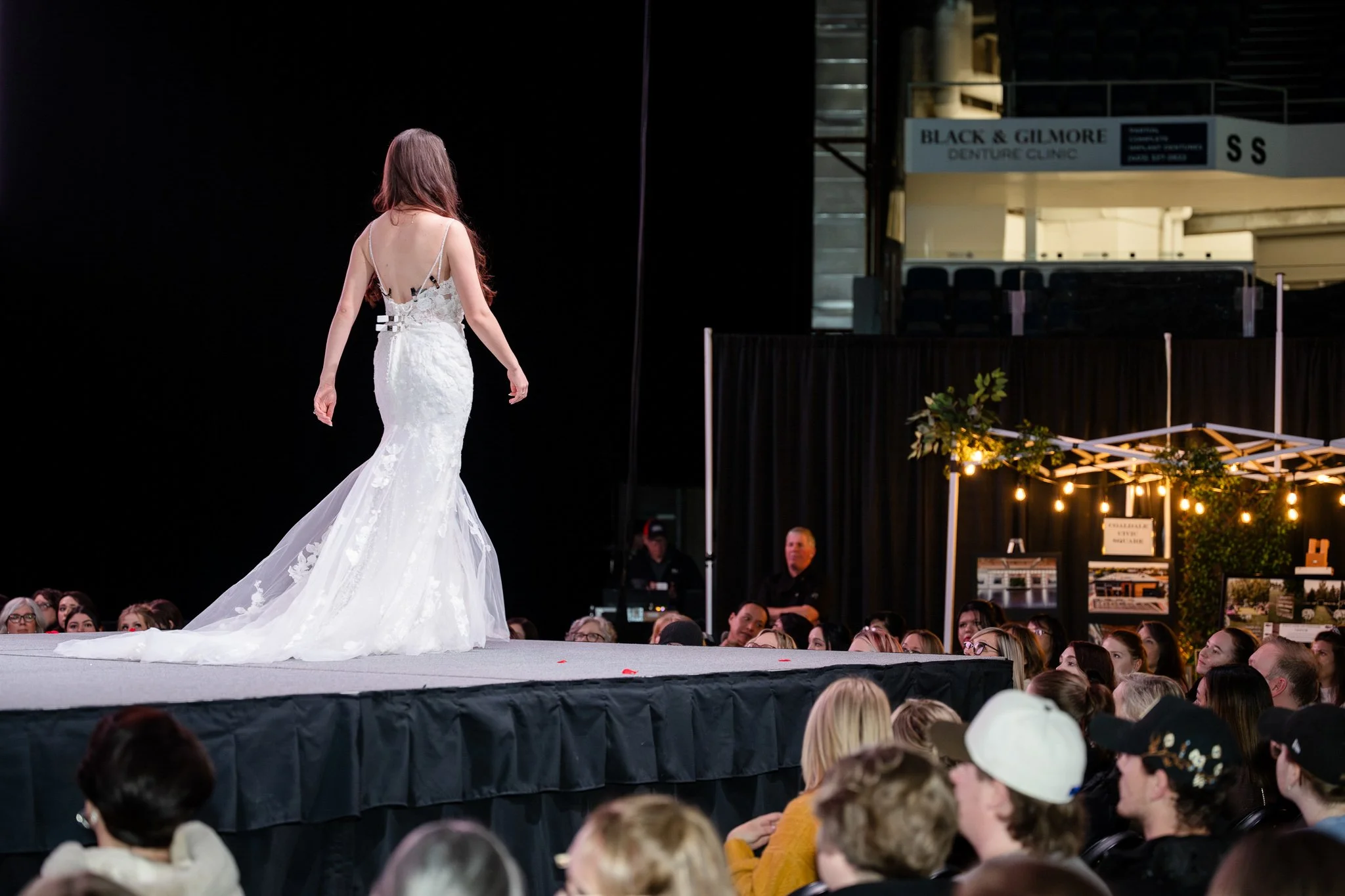 A woman in a white wedding dress walking on a runway during a fashion show, with an audience watching.