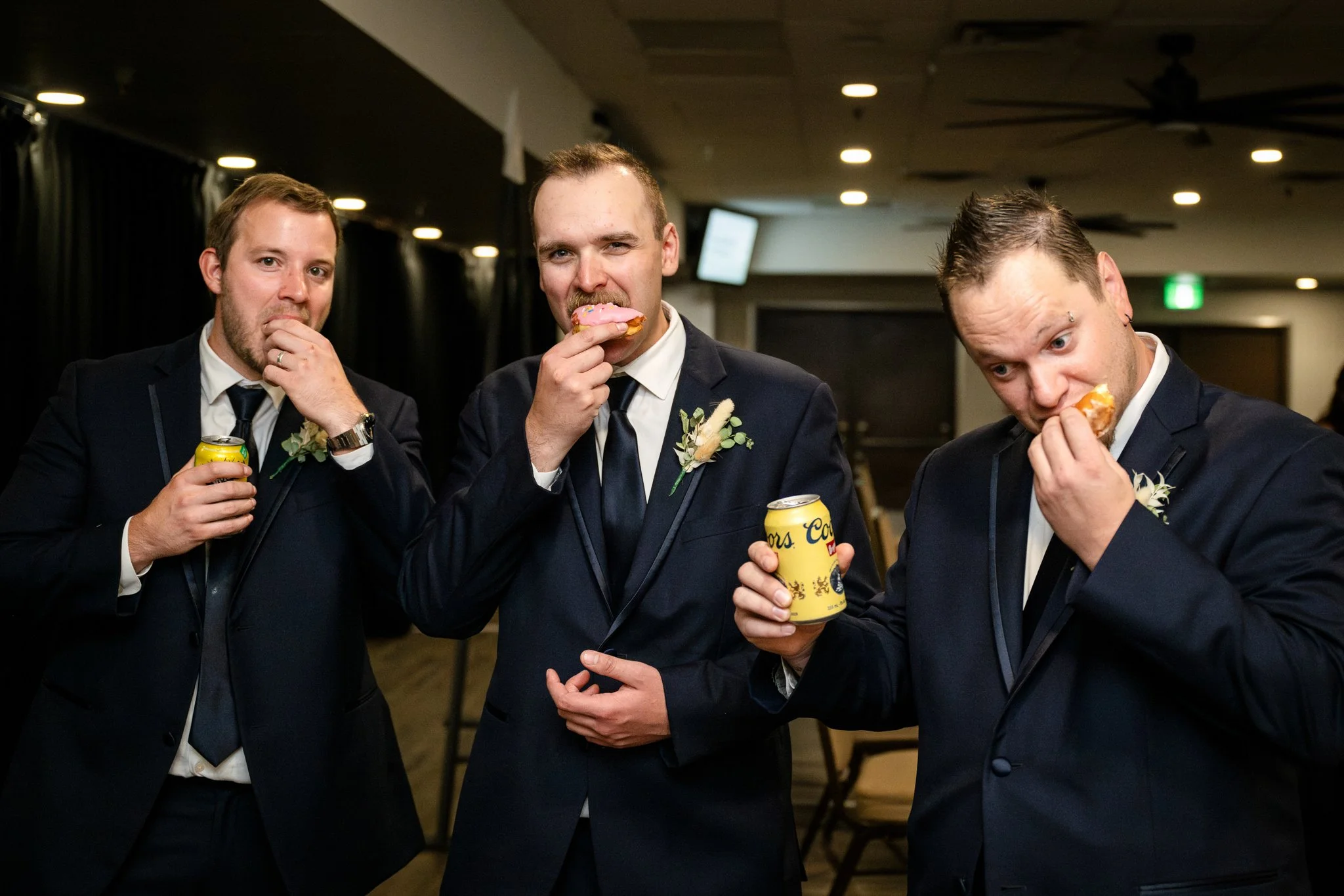 Three men in suits eating donuts and holding cans of beer at an indoor event.