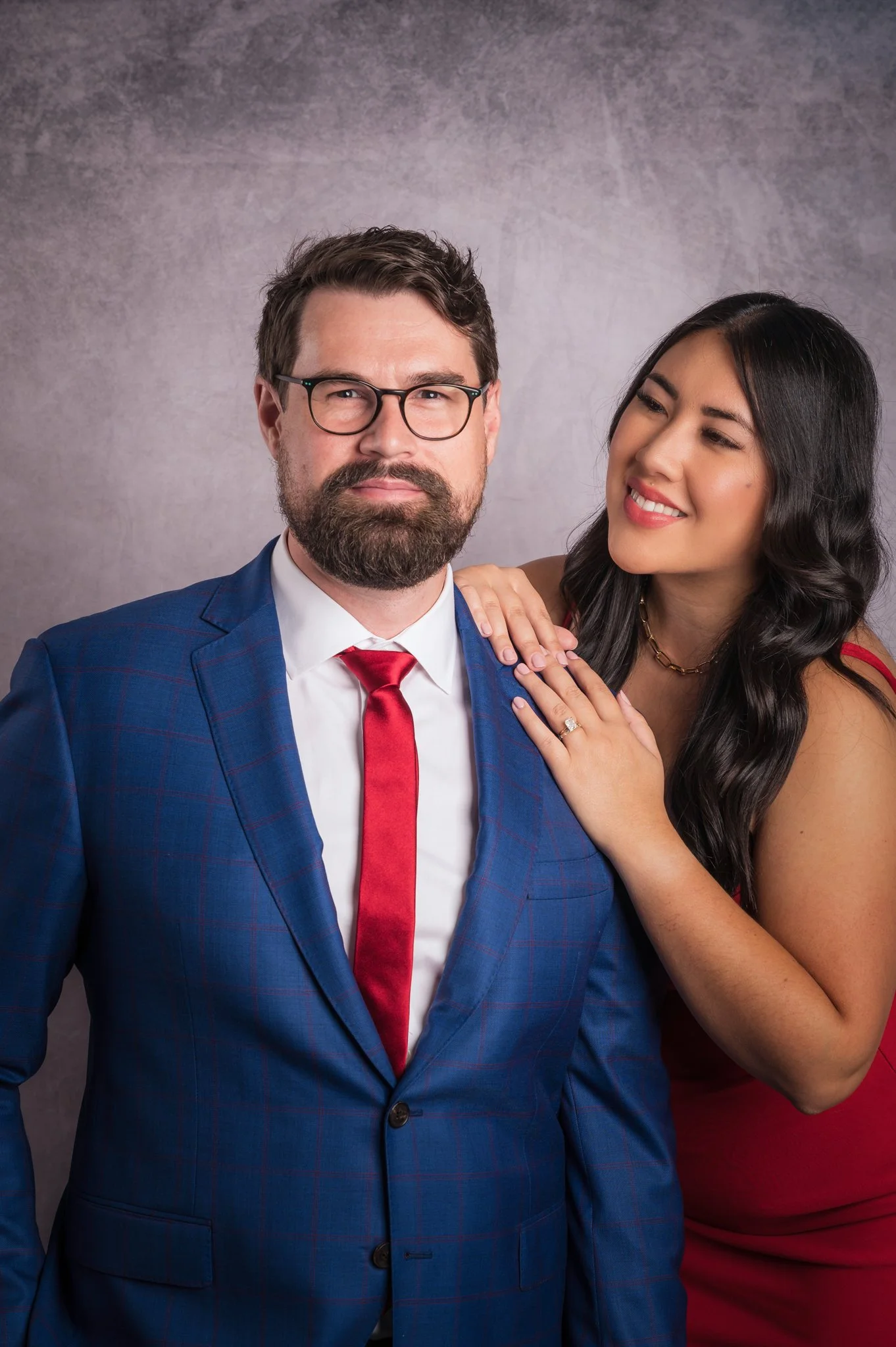 A man in a blue suit with a red tie and glasses standing next to a woman in a red dress, with the woman resting her hand on his shoulder and smiling at him.