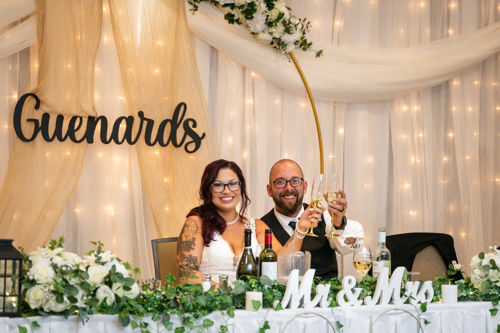 A newlywed couple sitting at a decorated wedding reception table, raising glasses for a toast, with a 'Guernards' sign and floral decorations in the background.