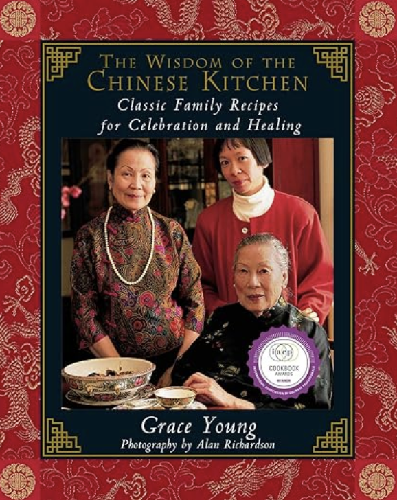 Book cover titled 'The Wisdom of the Chinese Kitchen: Classic Family Recipes for Celebration and Healing' by Grace Young, photograph by Alan Richardson. Features three women of Asian descent, two standing and one seated, in traditional attire, with Chinese dishes and bowls on the table in front of them.