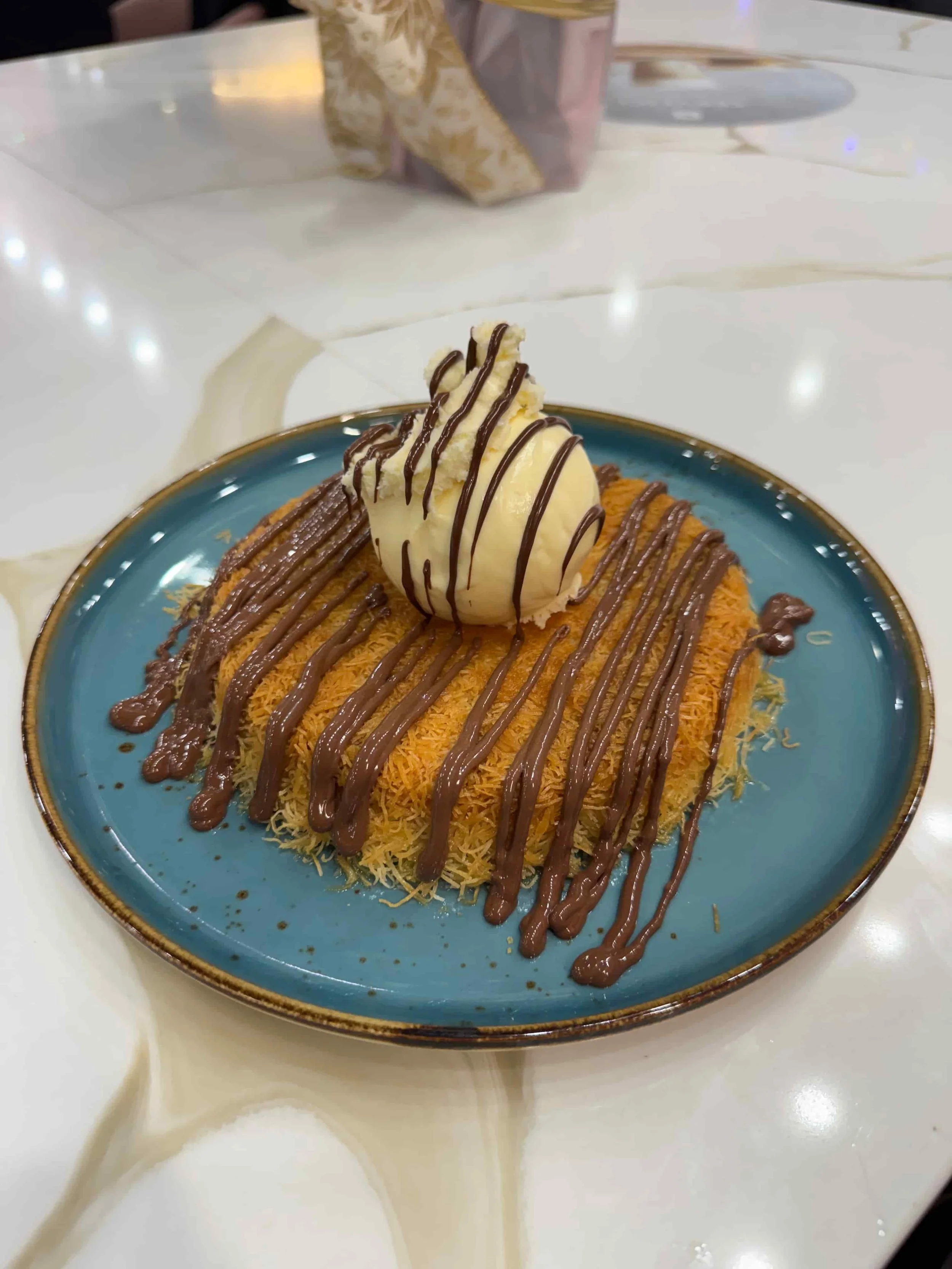 Dessert with shredded pastry, topped with a scoop of vanilla ice cream drizzled with chocolate sauce and served on a blue plate.