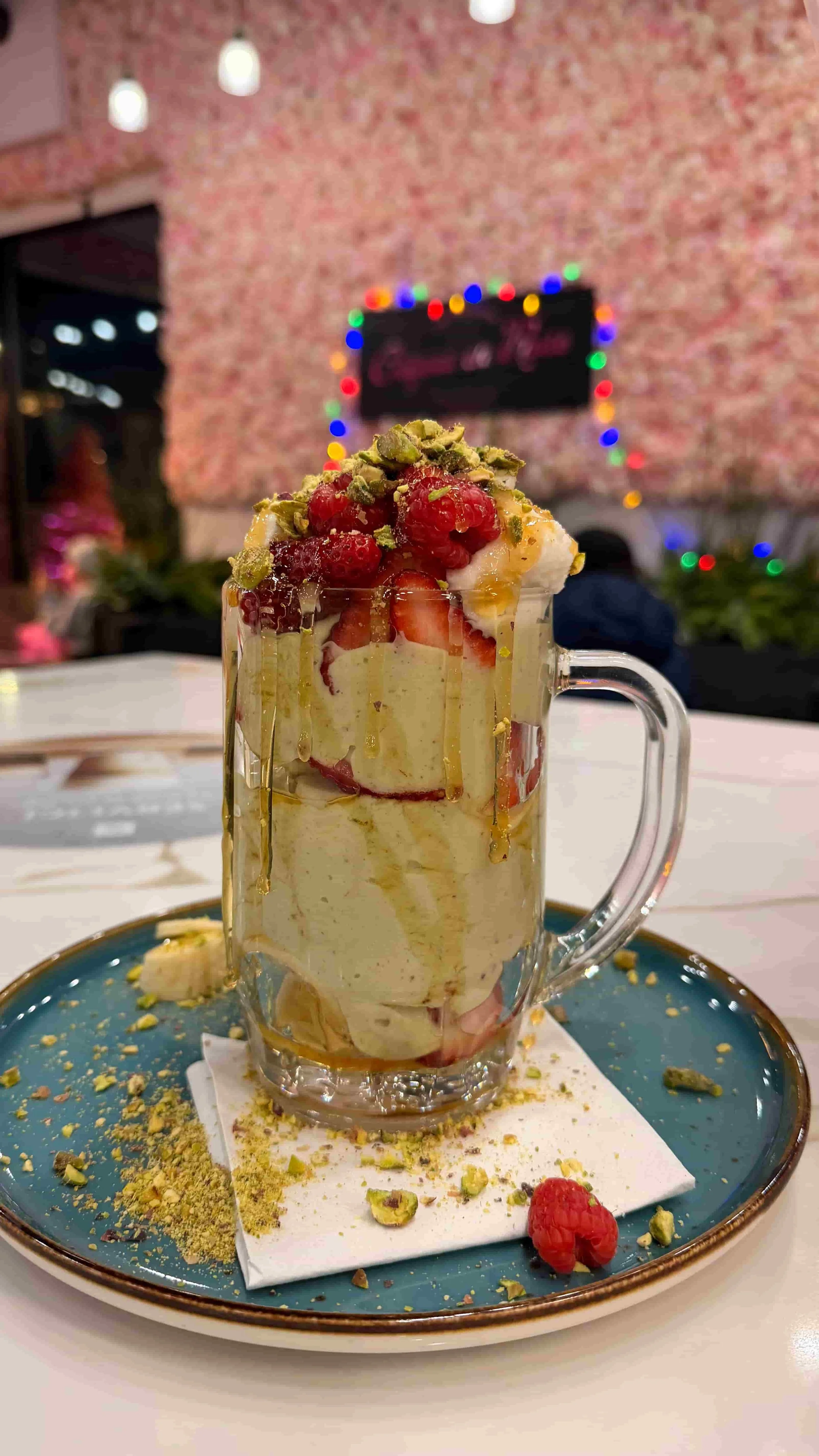 A tall glass mug of layered strawberry and cream dessert topped with raspberries, chopped pistachios, and drizzled with honey or syrup, set on a blue plate with crushed pistachios and a raspberry, in a cozy restaurant with colorful festive lights and