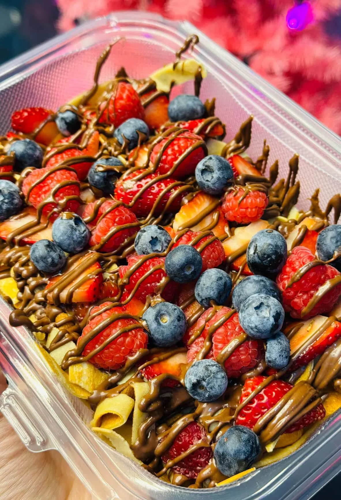 A fruit dessert in a plastic container topped with strawberries, blueberries, raspberries, and drizzled with chocolate.