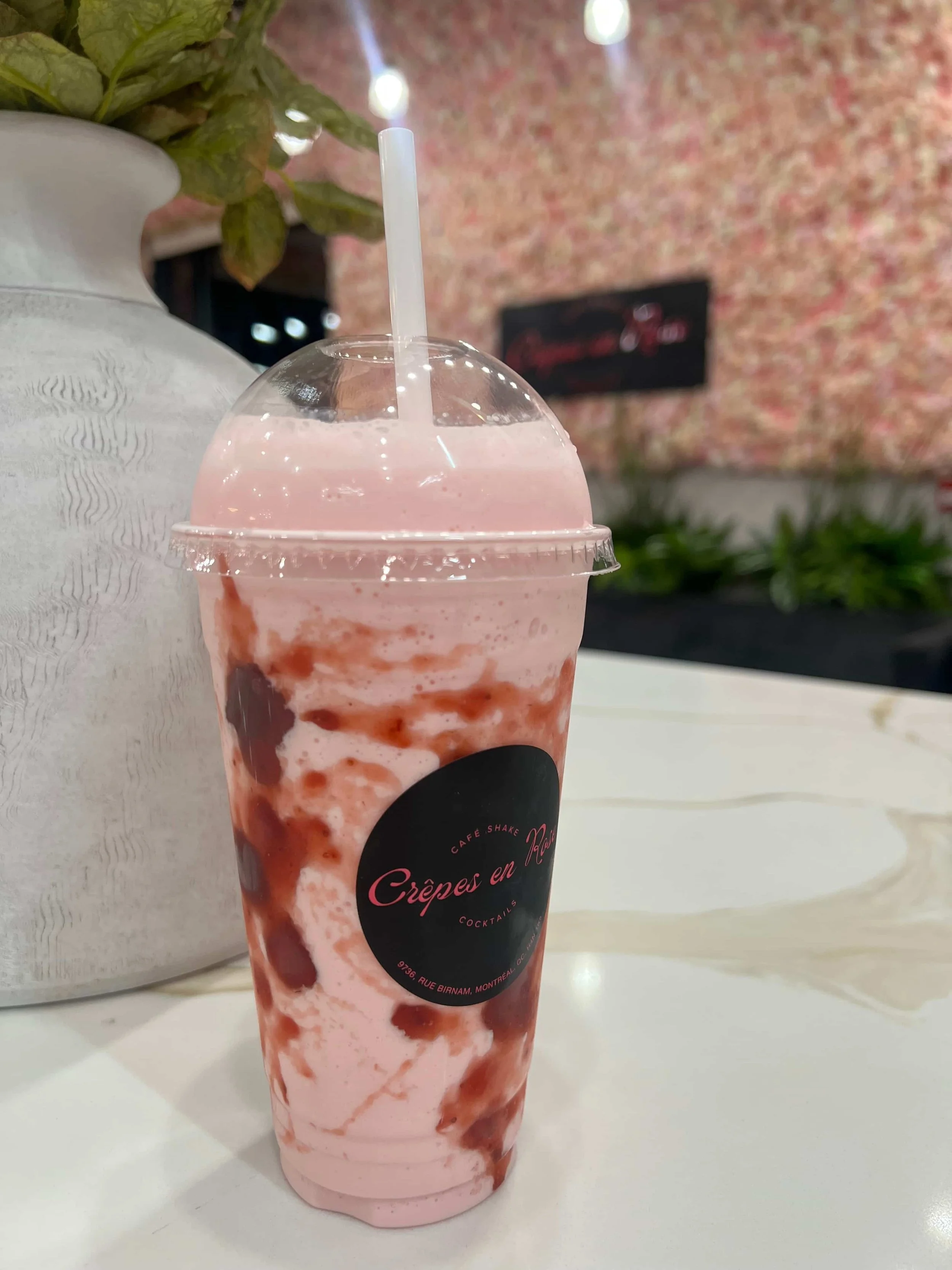 A pink and brown swirled milkshake in a clear plastic cup with a black label reading "Crépes en Rage" and a white straw, placed on a white table inside a cafe.