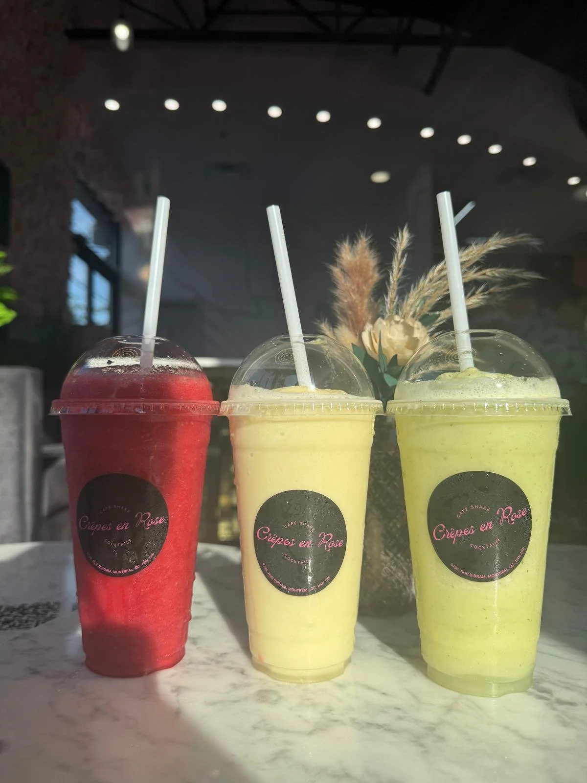 Three colorful smoothies in clear plastic cups with straws, placed on a white marble table. The cups have black labels reading 'Crêpes en Rose.' The smoothies are red, yellow, and green, with a background of a flower arrangement and a modern indoor s
