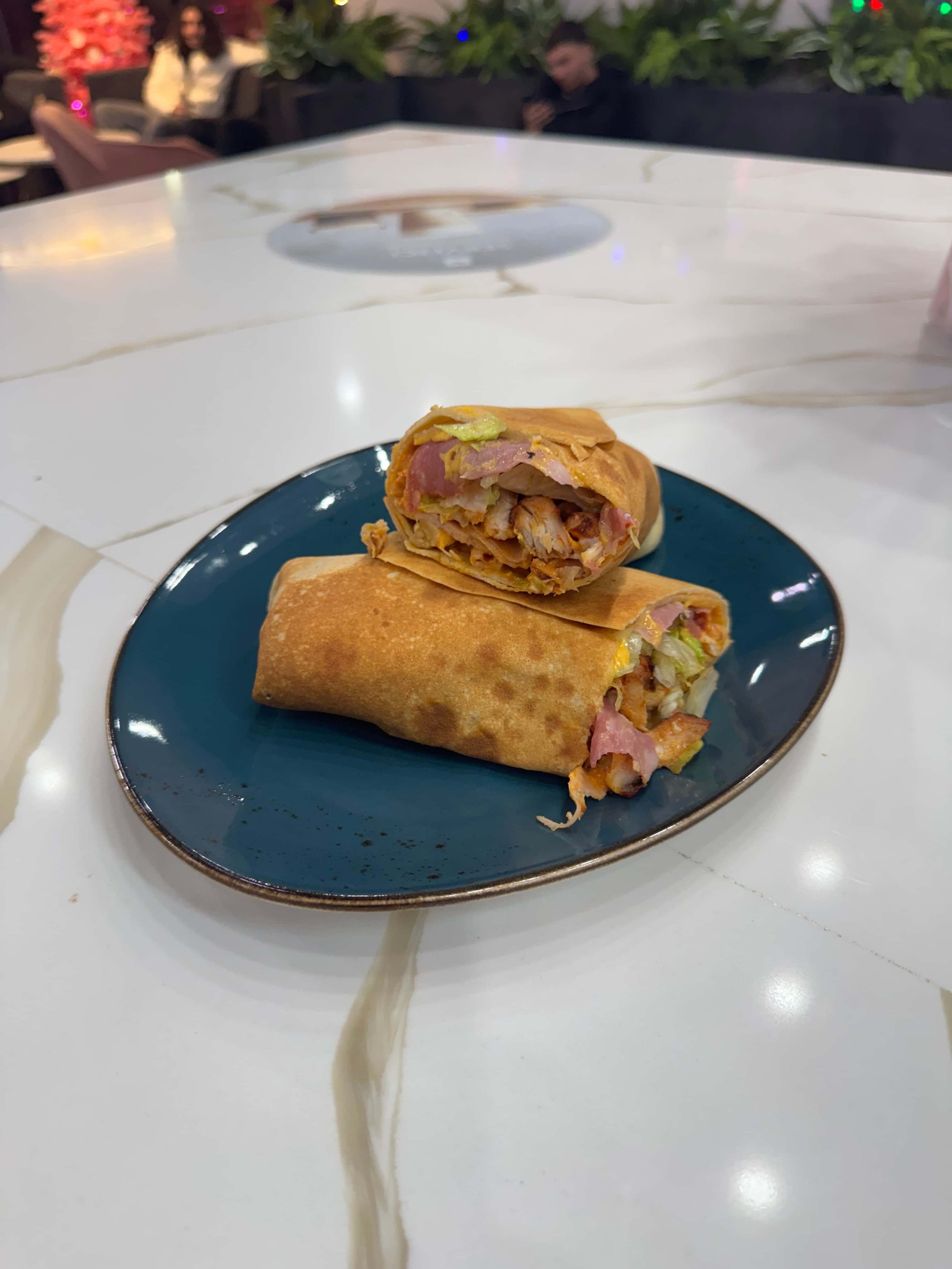 A spring roll sliced in half on a blue plate, revealing mixed vegetables, meat, and noodles inside, placed on a white marble table with people in the background.