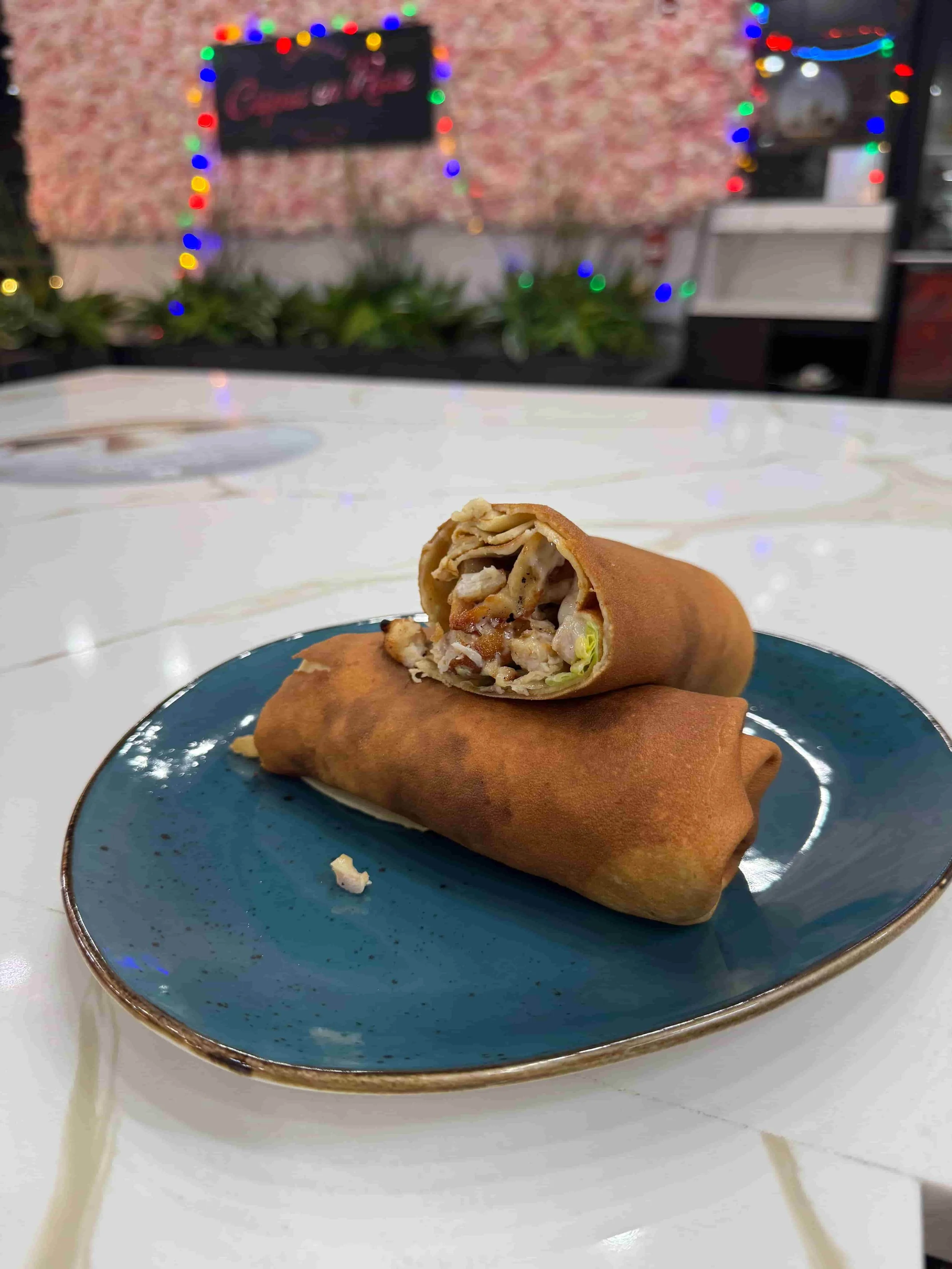 Two spring rolls on a blue plate, one cut open to show filling of chicken and vegetables, on a white marble table with blurred Christmas lights in the background.