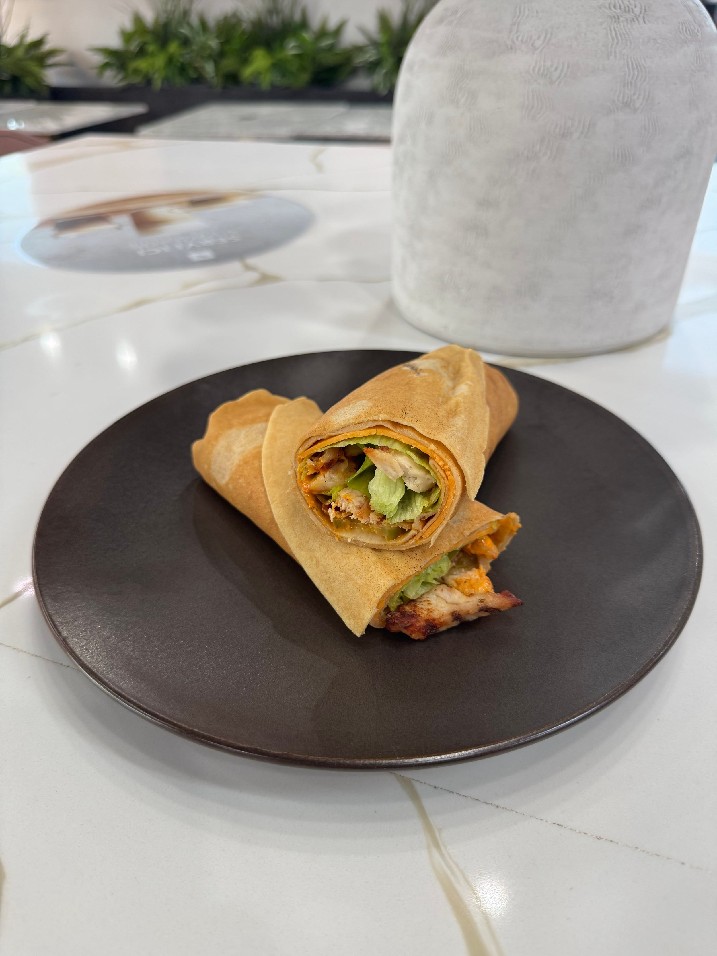 Two chicken wrap sandwiches on a black plate, with one sliced open revealing lettuce, chicken, and other ingredients, on a white table with a gray cup and a blurred outdoor background.