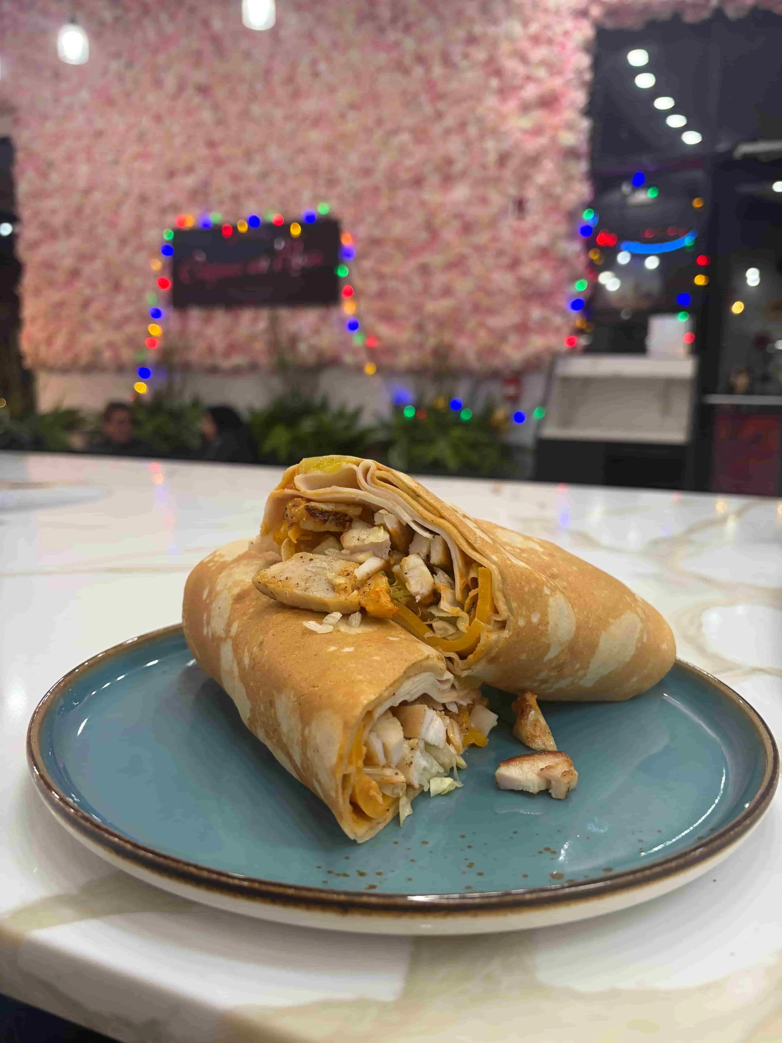 Two chicken wrap sandwiches on a blue plate with a blurred background of a restaurant decorated with colorful Christmas lights.
