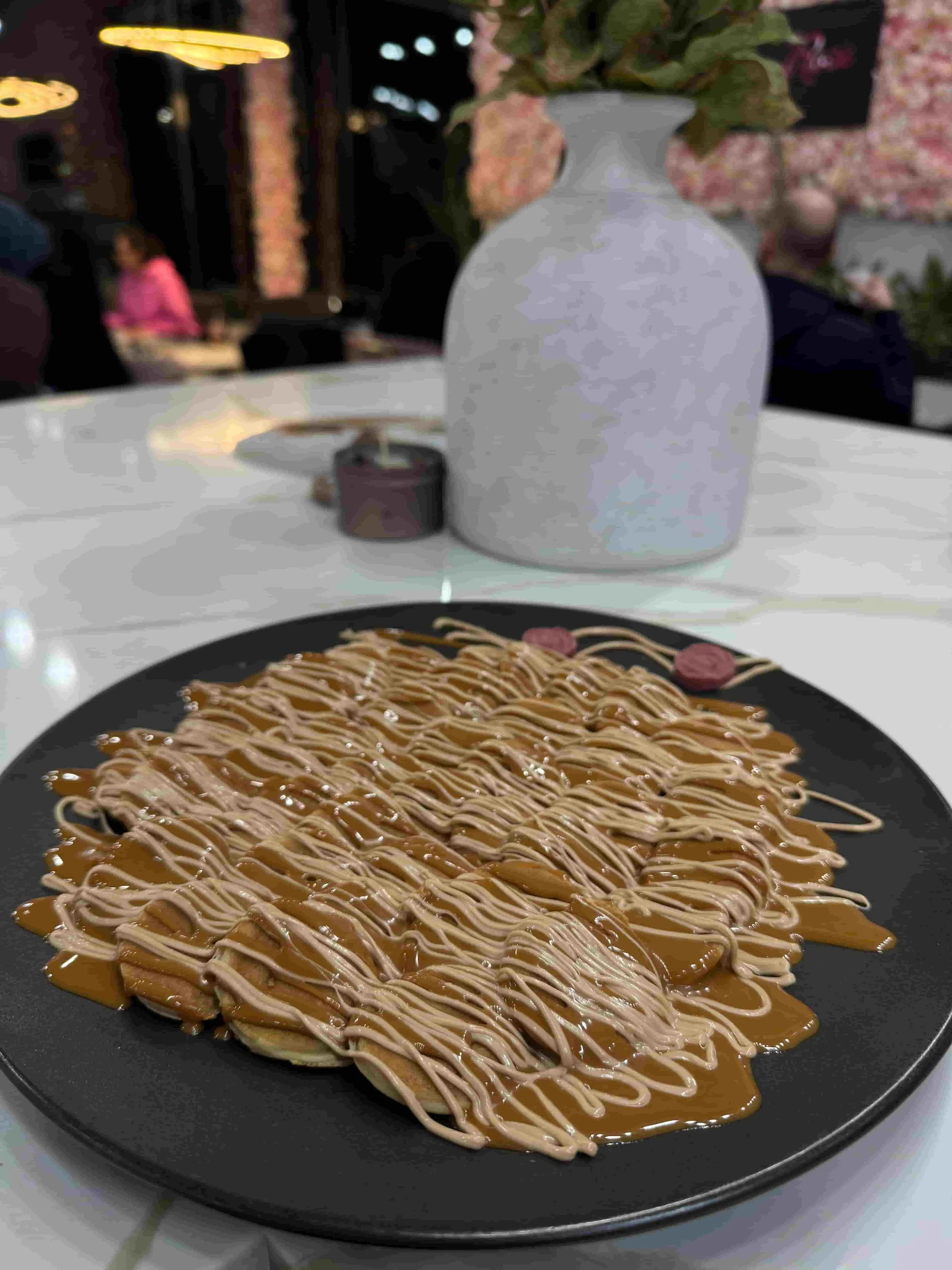 Plate of dessert with caramel and white drizzle over cookies on a table indoors, with a white vase, a small candle, and a pink flower arrangement in the background.
