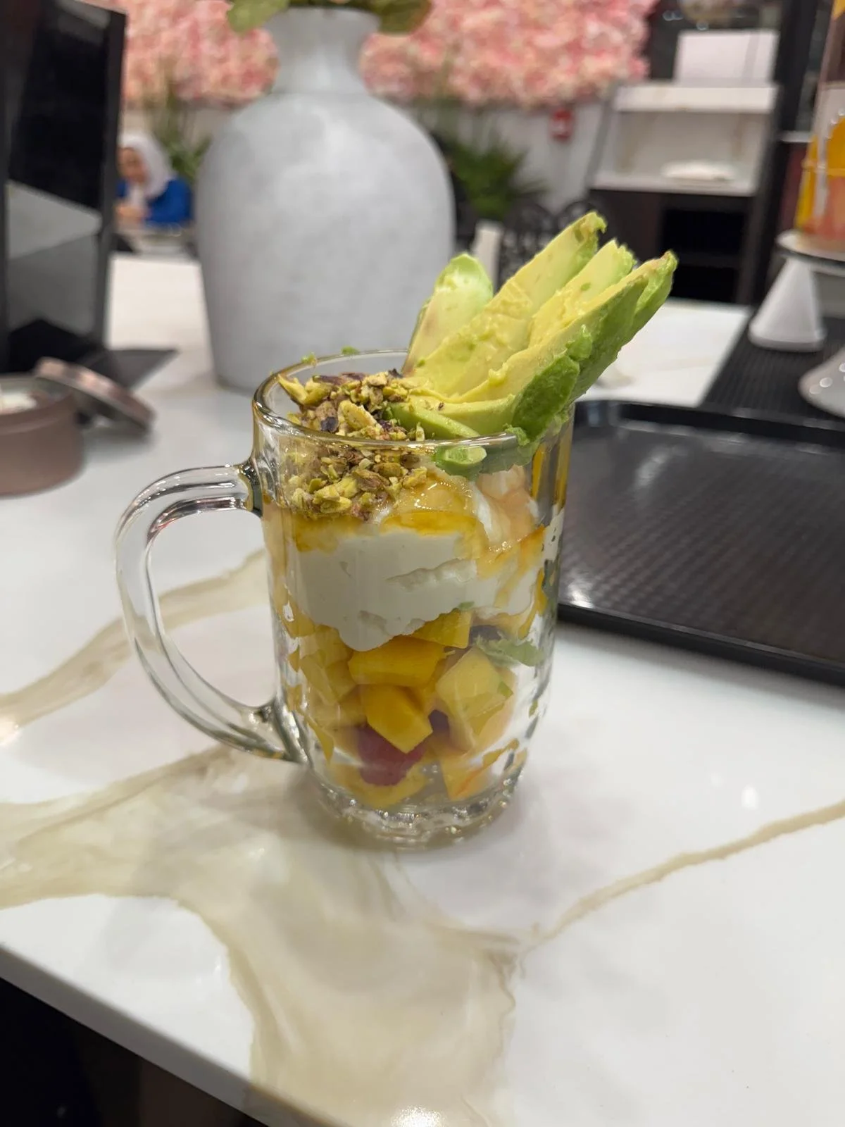 Mango, avocado, and yogurt parfait topped with chopped pistachios in a clear mug on a white table.