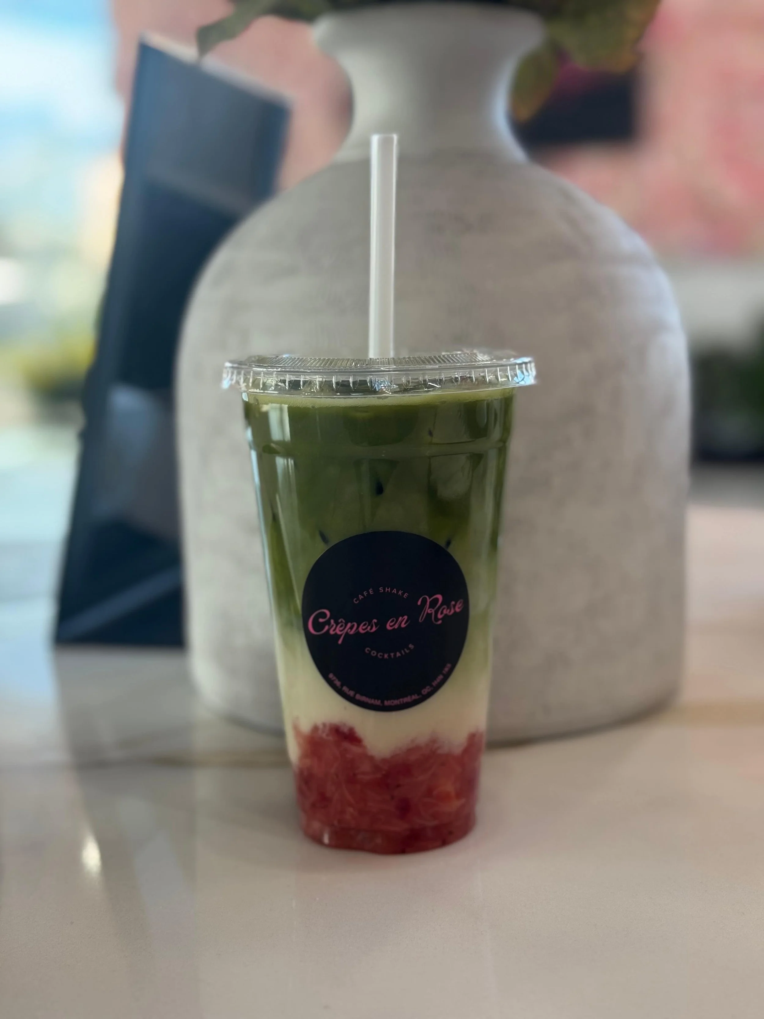 A clear plastic cup with a green and cream layered smoothie topped with red fruit pieces, strawberry syrup at the bottom, labeled "Crêpes en Rose" with a black background and pink text, with a straw, placed on a light-colored table with a large white