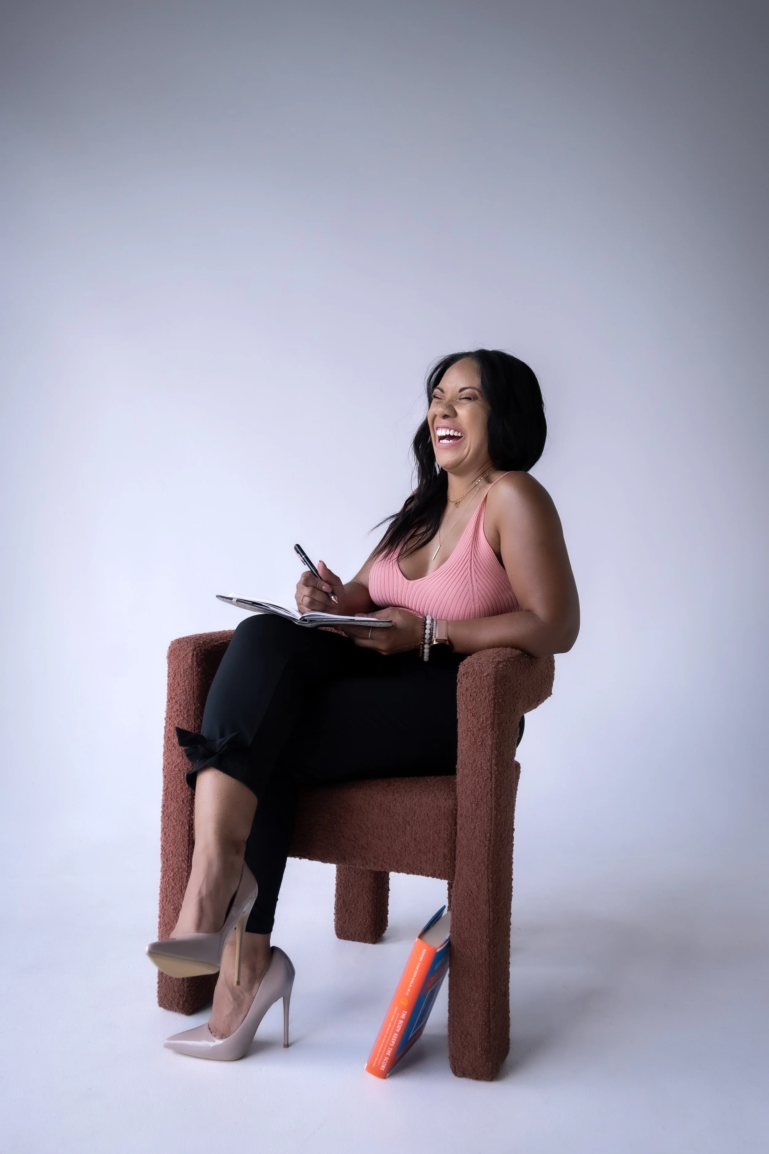Woman sitting in a chair, laughing, writing in a notebook, wearing a pink top and black pants with beige high heels. A couple of books are under the chair's leg.