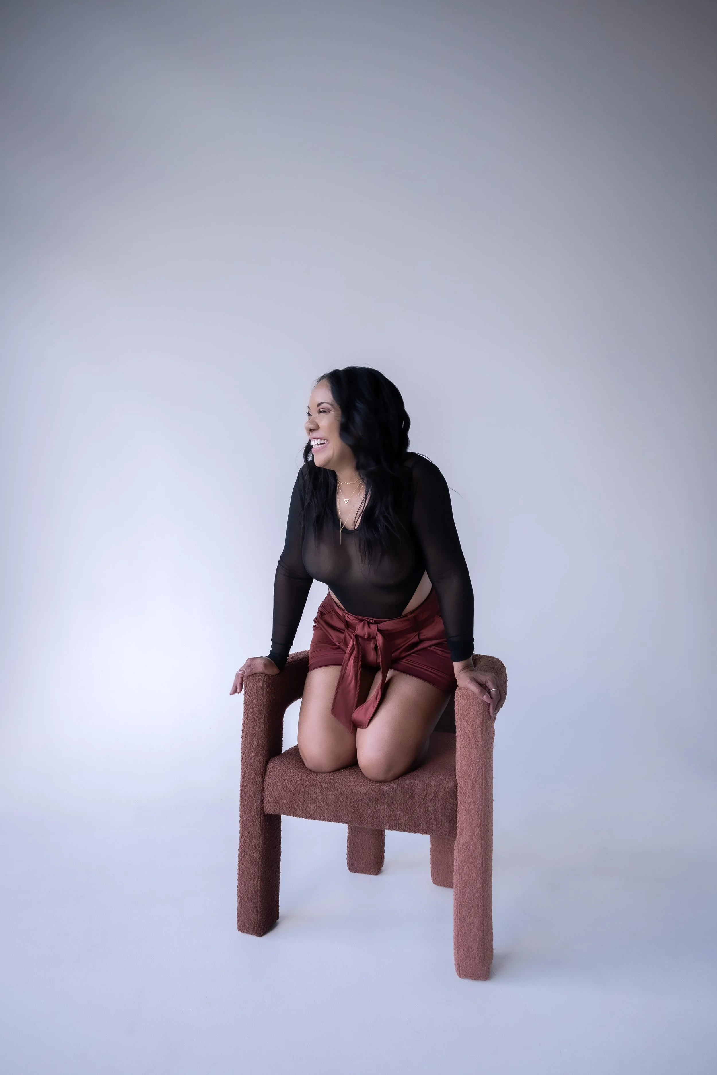 Woman in a black top and red skirt kneeling on a modern chair against a plain background, smiling.