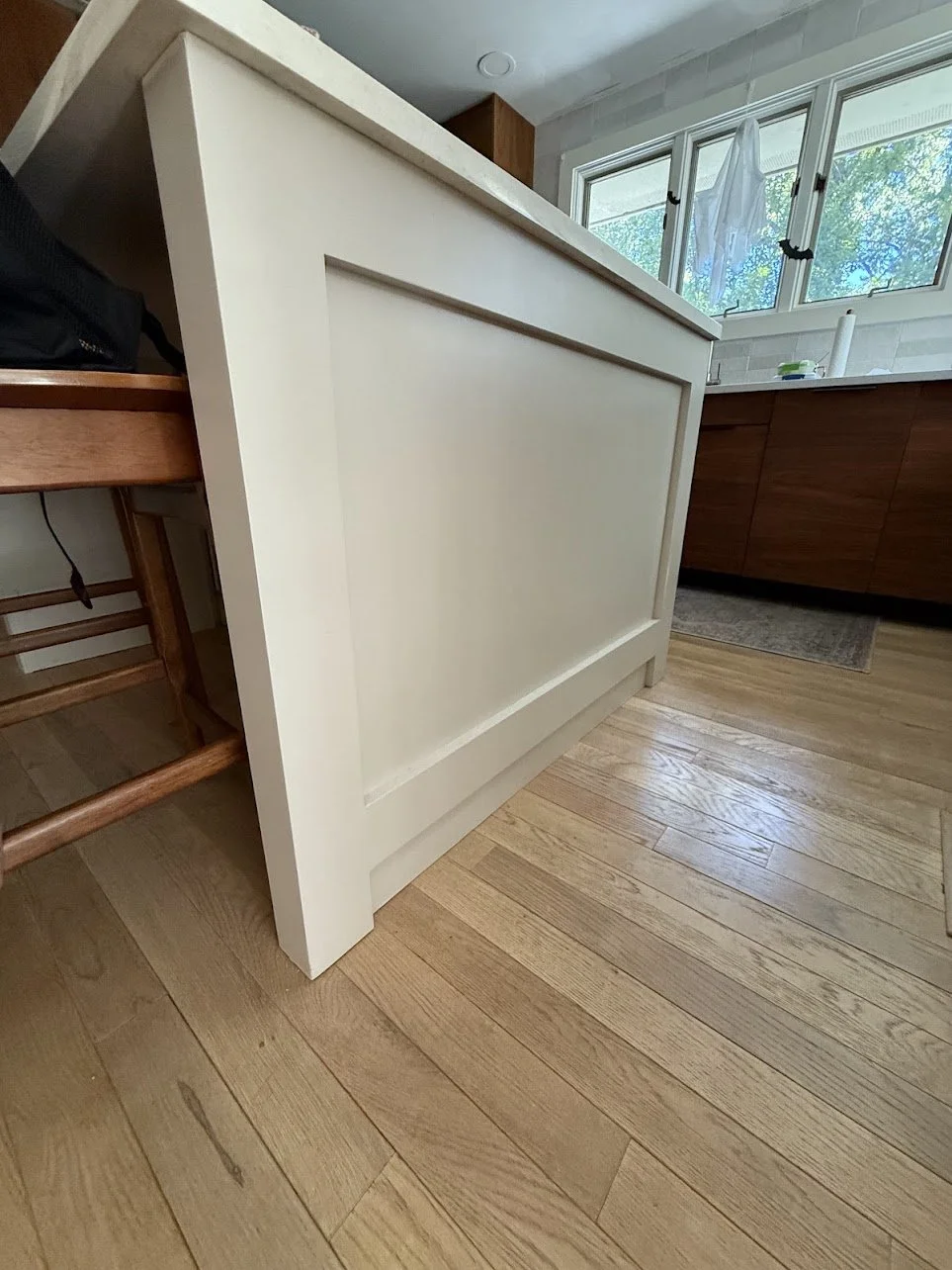 Close-up of a white kitchen island or counter with wooden cabinets in the background, in a bright kitchen with a window showing outdoors.