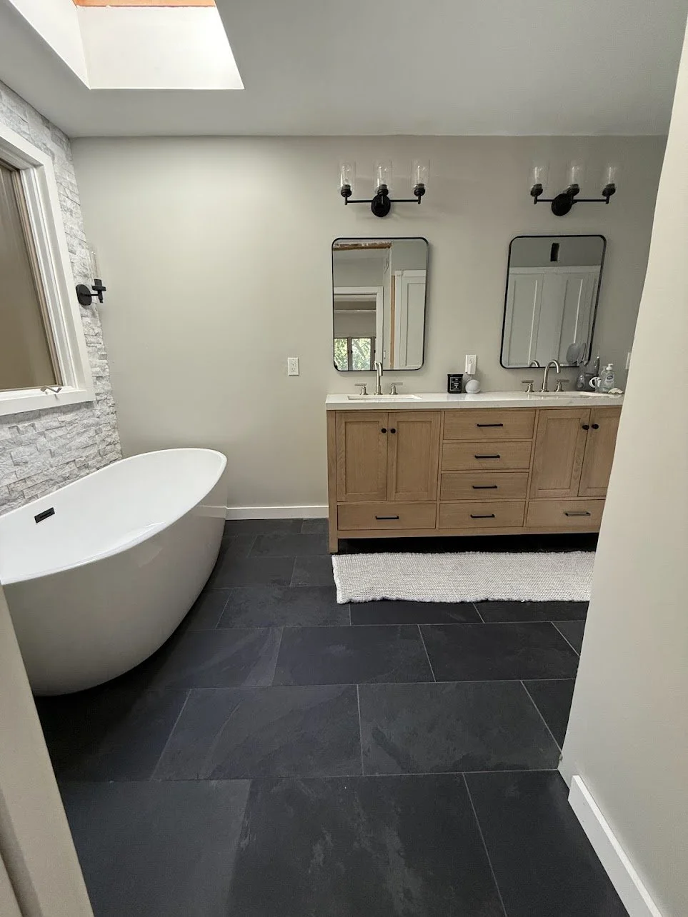 A modern bathroom with a white freestanding bathtub, a wooden vanity with two sinks, two mirrors, black light fixtures, and a skylight.