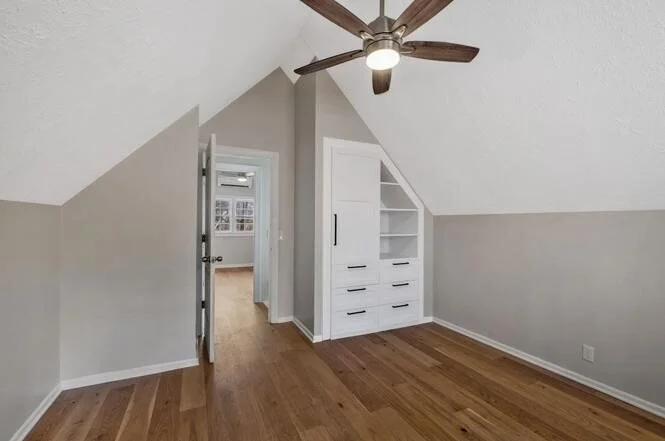 Empty room with sloped ceiling, built-in white bookshelf, wooden floor, ceiling fan, and doorway leading to another room.