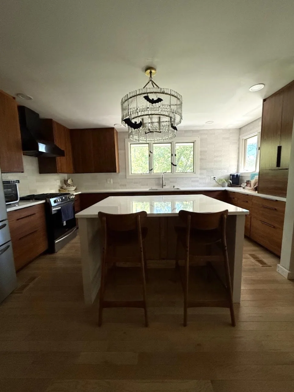 A modern kitchen with wooden cabinets, a white kitchen island, and a chandelier decorated with bats hanging from the ceiling. There are windows showing outside greenery.