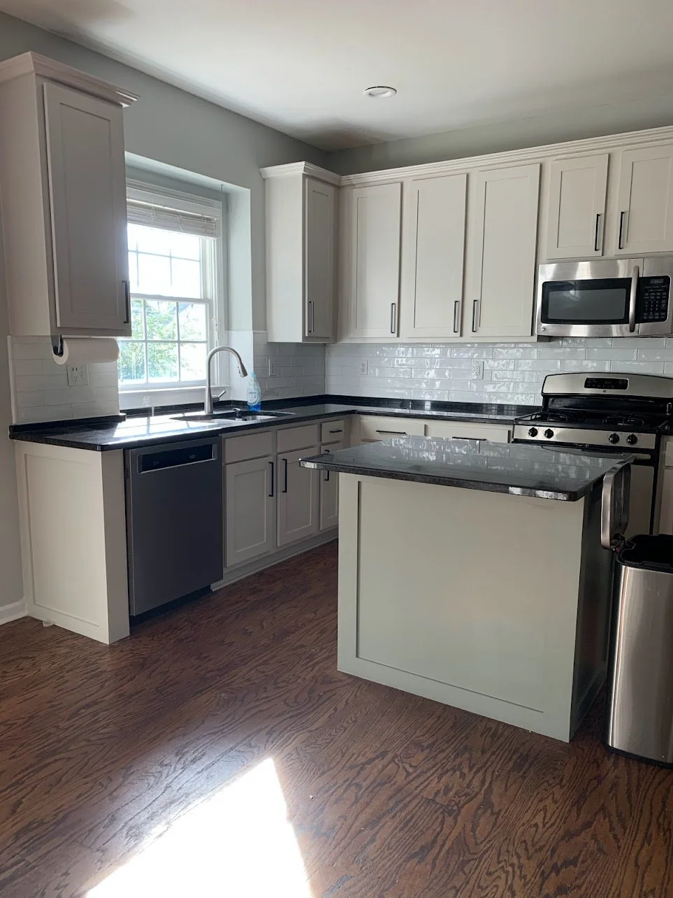 Modern kitchen with white cabinets, black countertops, stainless steel appliances, wooden floor, and a window above the sink.