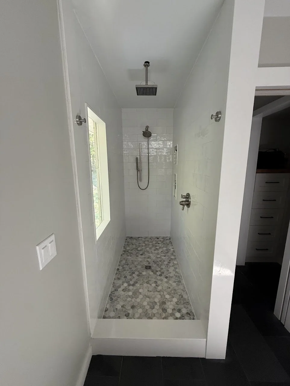 Empty walk-in shower with pebble tile floor, white tiled walls, a mounted showerhead, a handheld showerhandle, and a small window on the left wall.