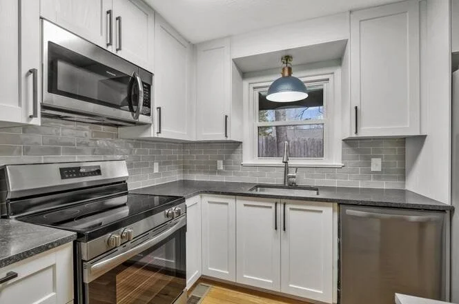 Modern kitchen with white cabinets, gray countertop, stainless steel appliances including a microwave and stove, a gray subway tile backsplash, a window above the sink, and a blue pendant light above the window.