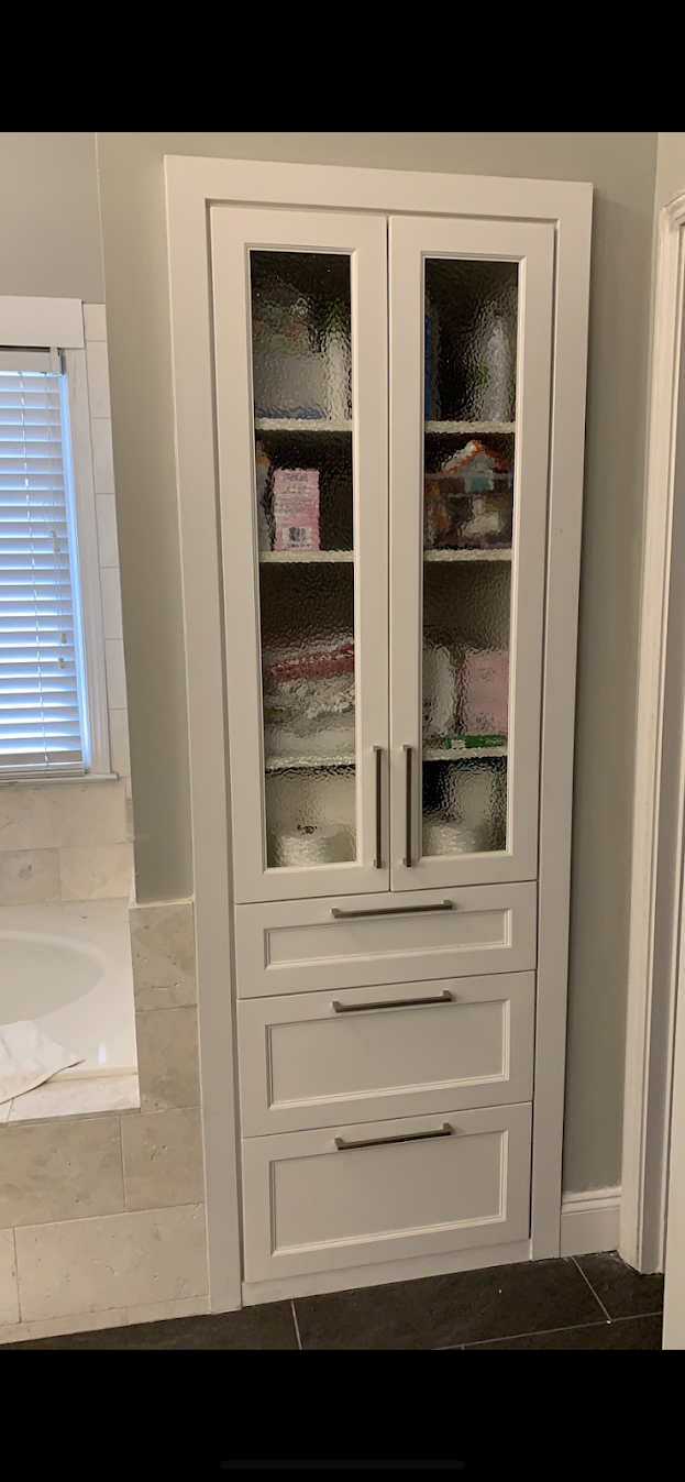 A white bathroom cabinet with frosted glass doors and four drawers below, situated next to a bathtub with a towel inside.