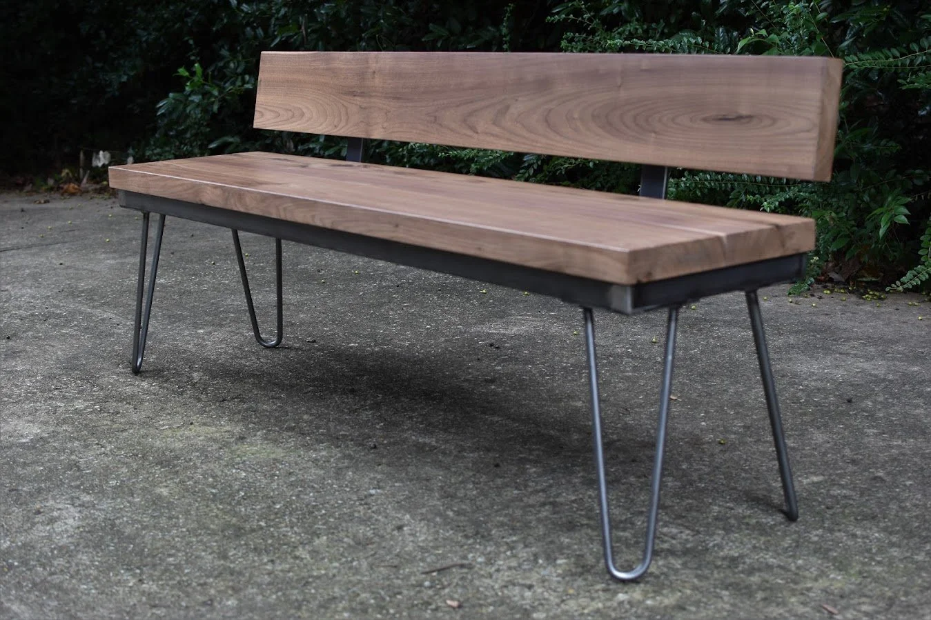 A wooden bench with metal legs on a concrete surface, with greenery in the background.