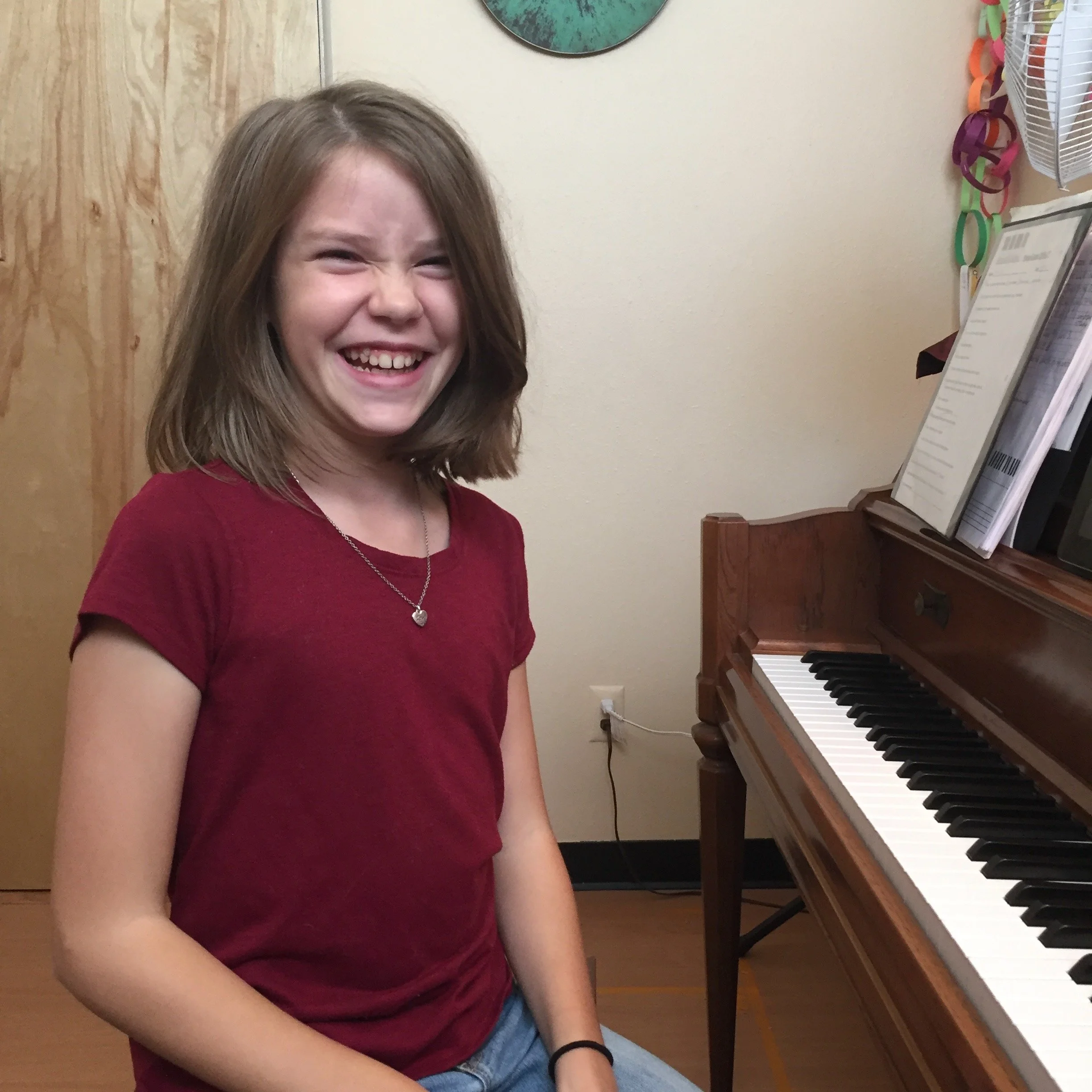 A girl with shoulder-length brown hair wearing a red t-shirt and a necklace with a heart pendant, smiling and sitting near a piano with sheet music on top.