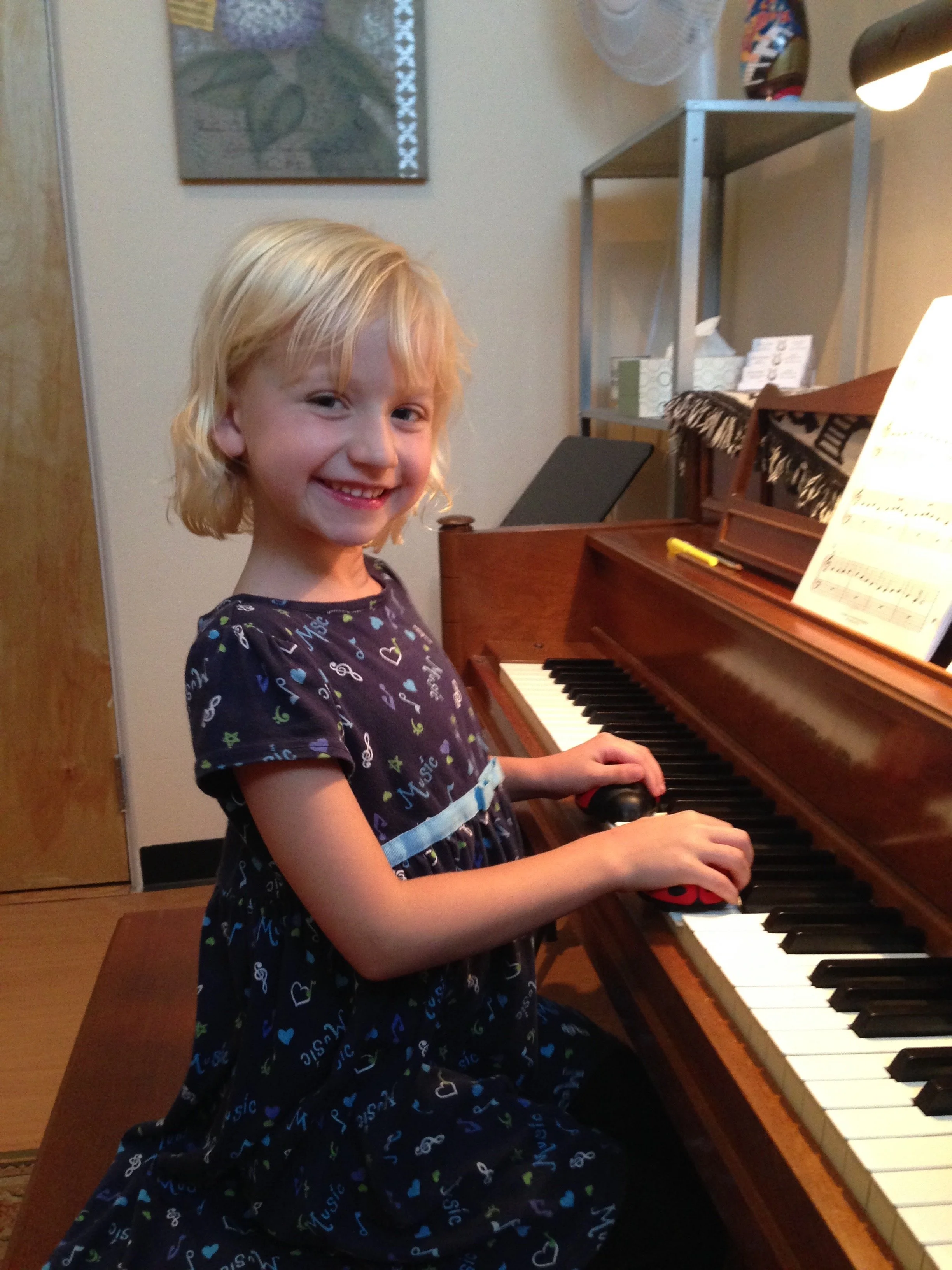A young girl with blonde hair sitting at a piano, smiling at the camera, with sheet music on the stand.
