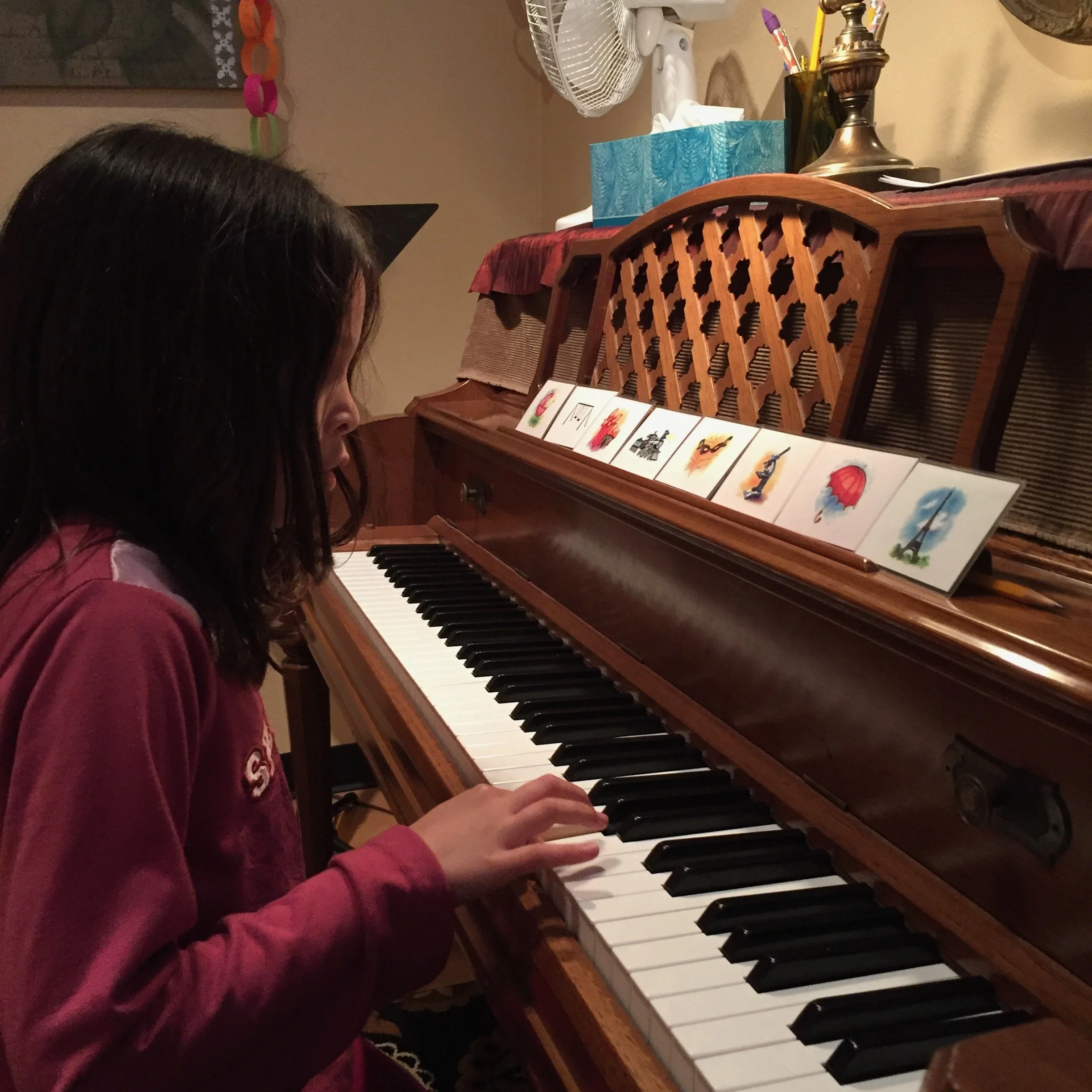 A young girl with dark hair playing an upright piano with picture cards on top, in a cozy room.