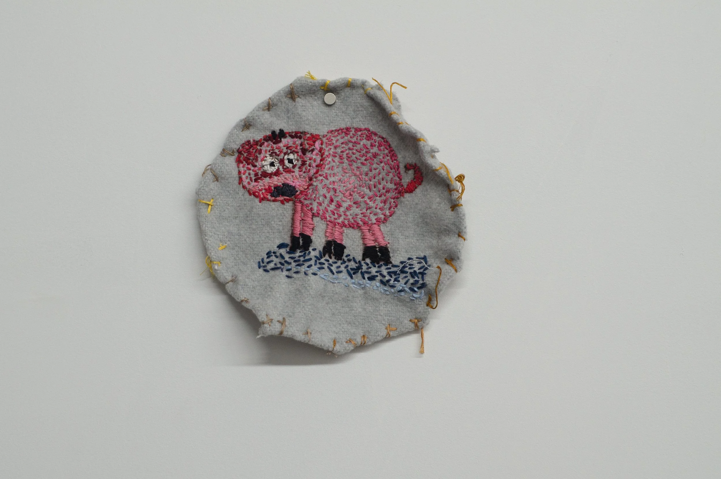 A round piece of knit fabric edged in loose yellow stitching. A pink hand embroidered pig stands upon a blue surface in the center.