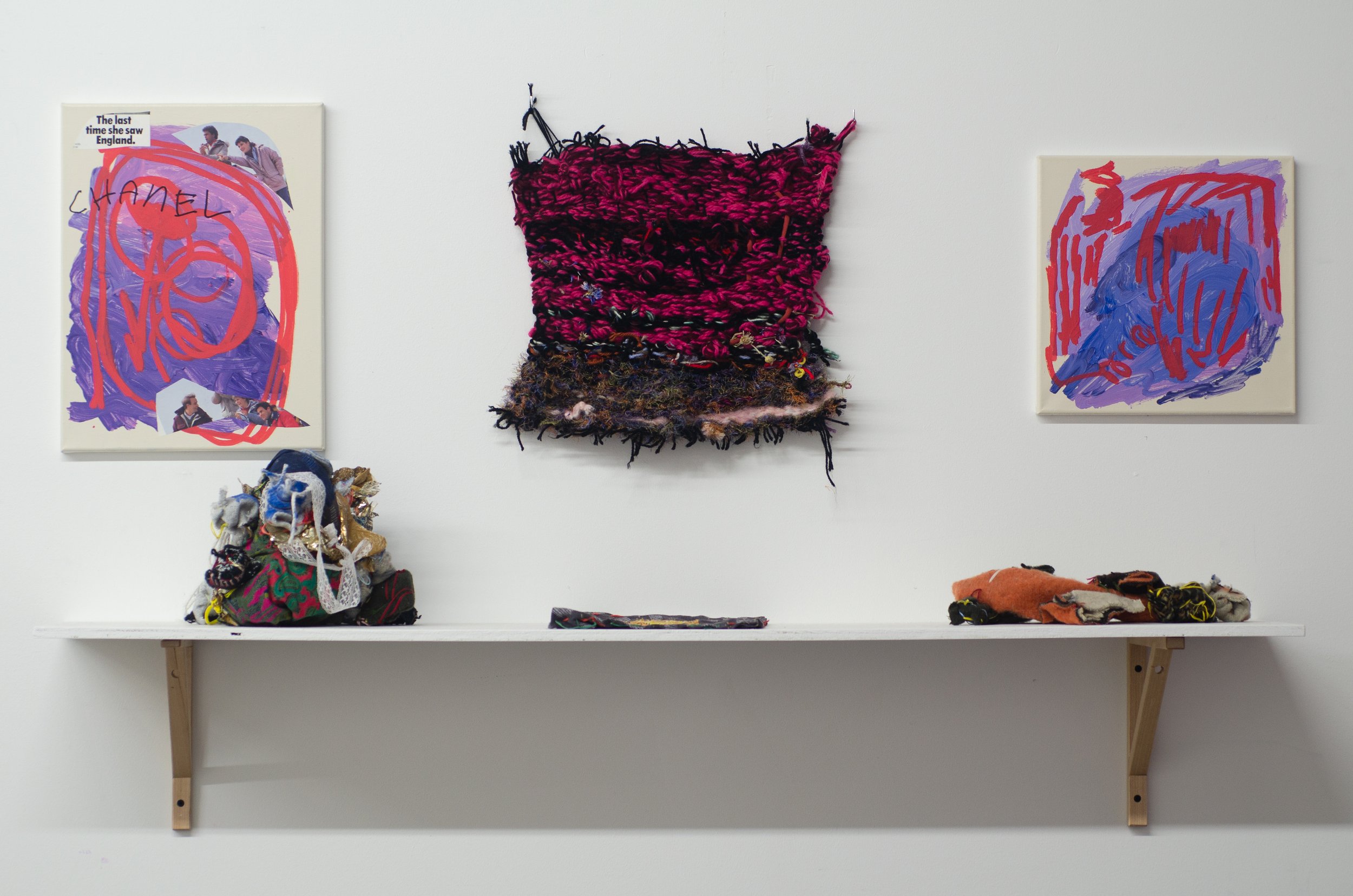 Installation view of a shelf supporting three soft sculptures beneath a mostly pink and black handmade weaving, and two canvases filled with loose brushstrokes in fuscia, red, and purpse, and found images of people