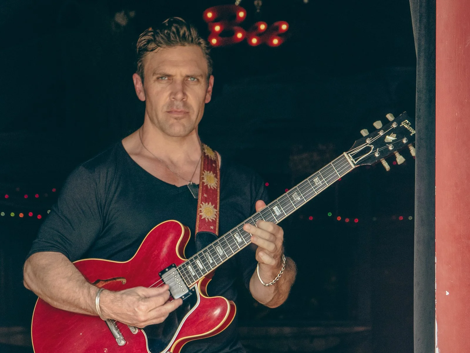 A man playing a red electric guitar in a dark setting with red and green string lights in the background.