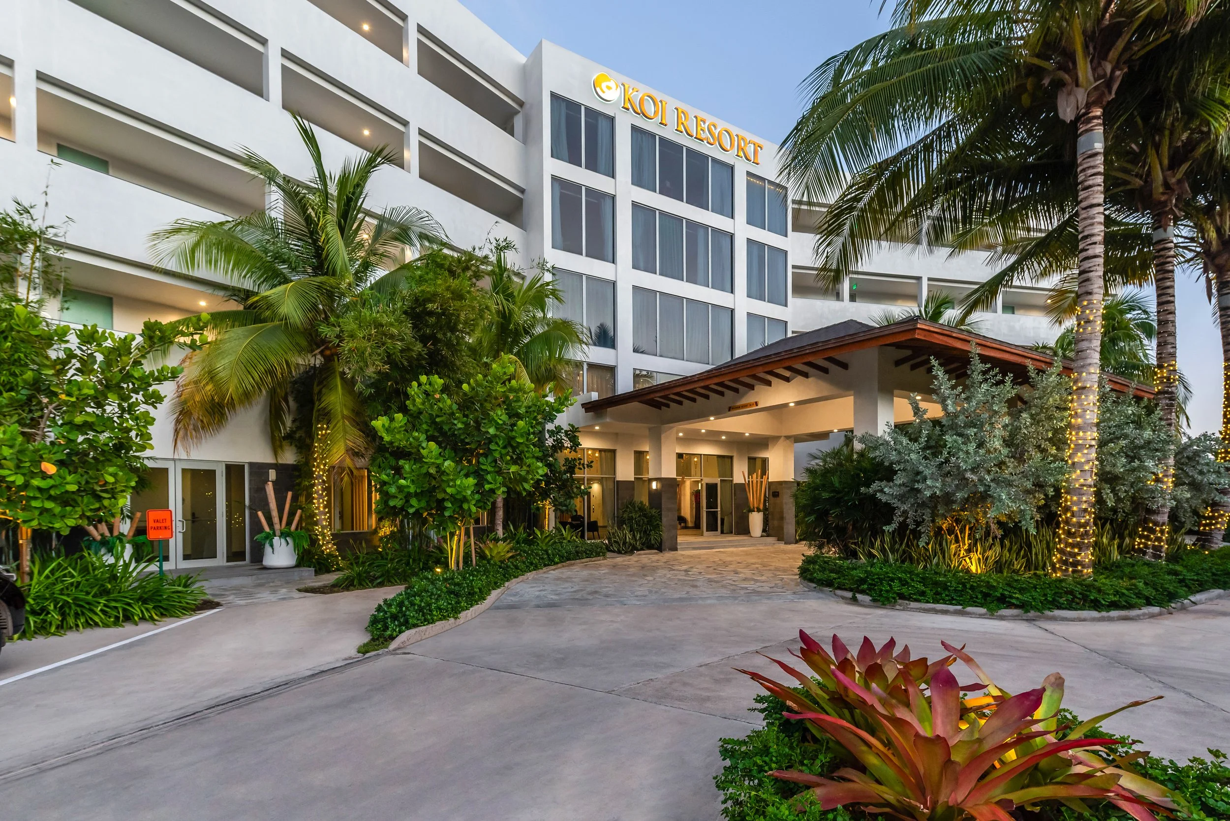 Exterior view of KOI Resort with tropical landscaping including palm trees and bushes, illuminated by evening lights, and a multi-story building with balconies and large glass windows.