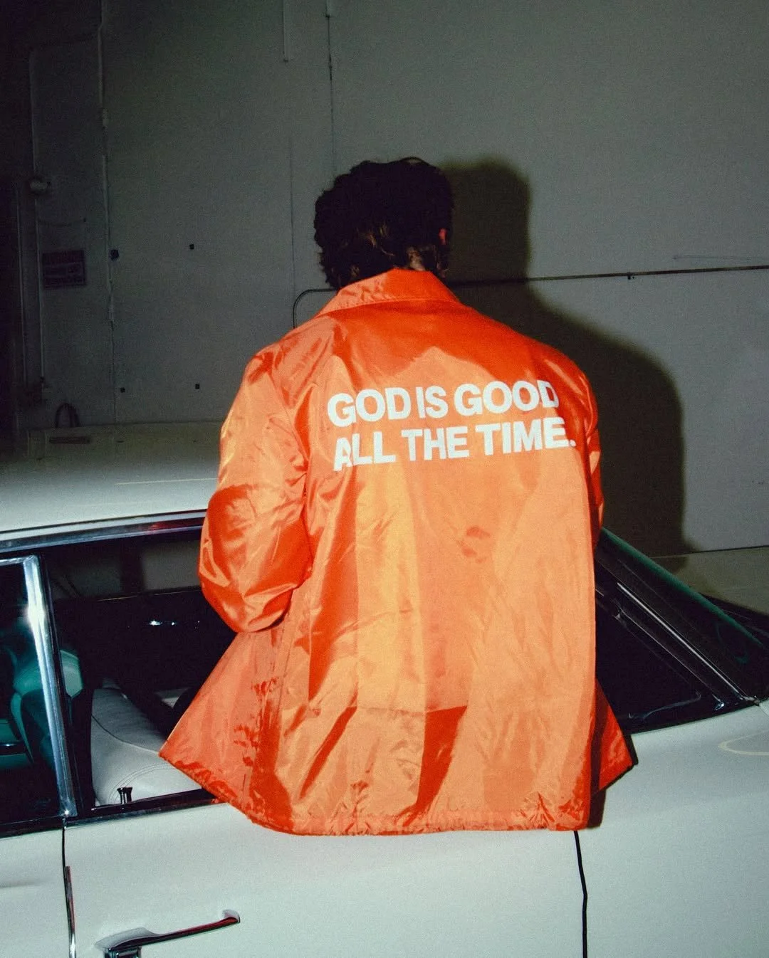 Person with dark, curly hair wearing an orange jacket with the text 'GOD IS GOOD ALL THE TIME' on the back, standing beside a white car in a garage or warehouse setting.