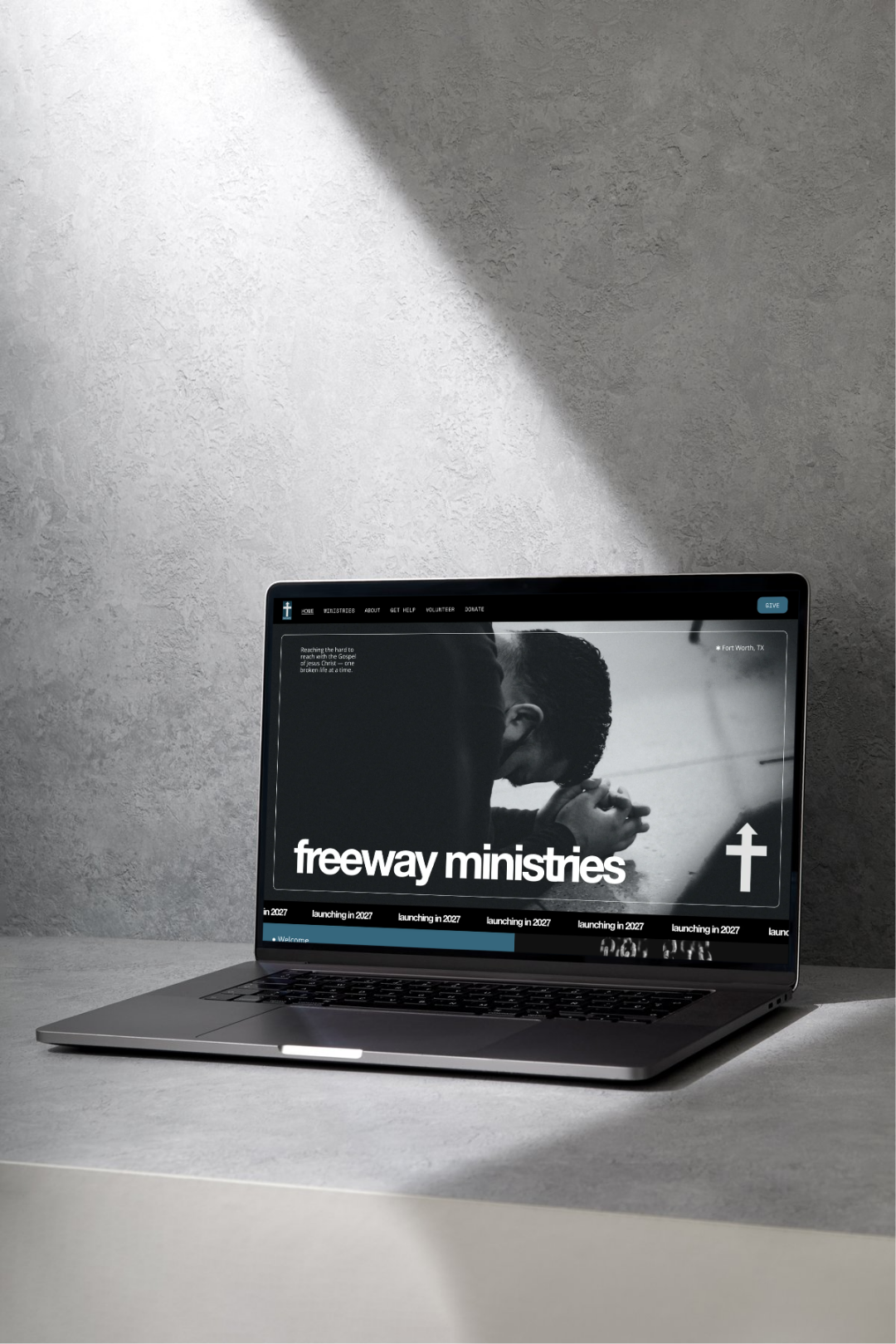 A laptop on a gray surface displaying a website for 'Freeway Ministries,' featuring a black and white image of a person praying or praying with their head bowed and hands clasped, with a cross symbol on the screen.
